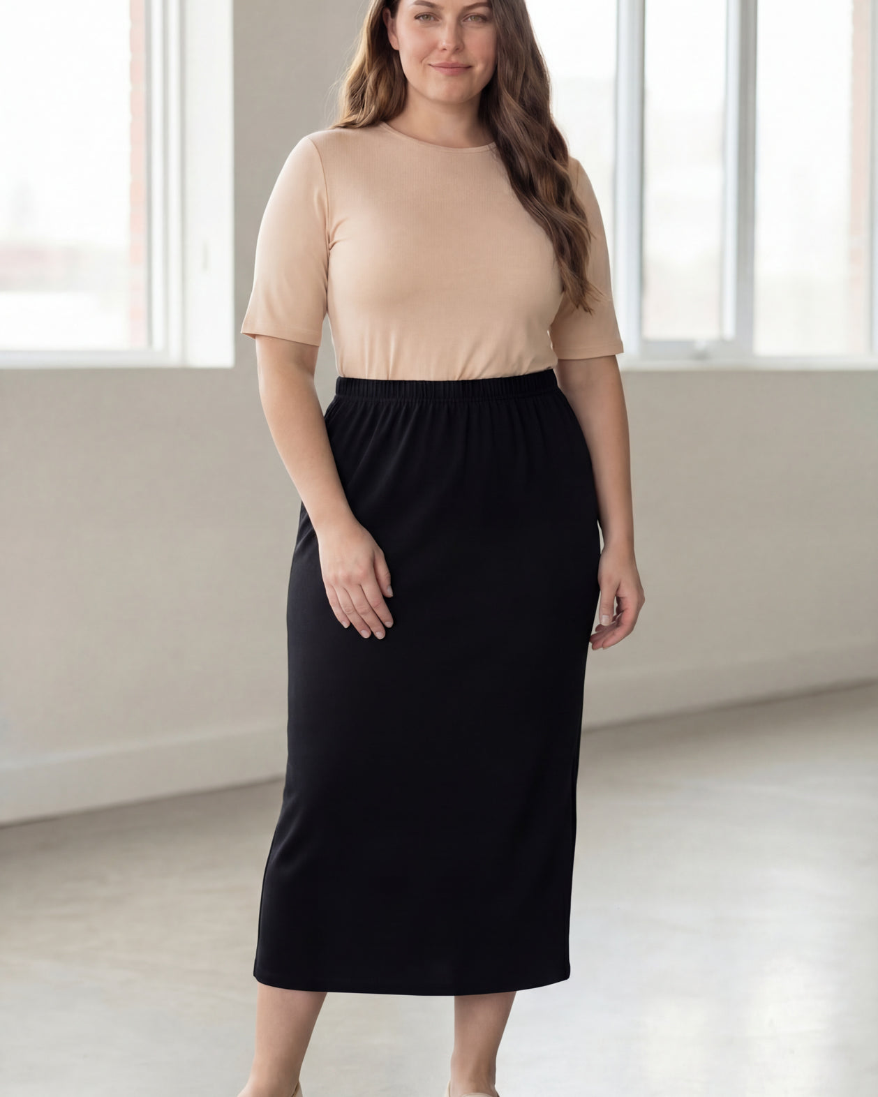 Woman wearing a beige shirt & black maxi length pencil skirt standing in a bright room with large windows.