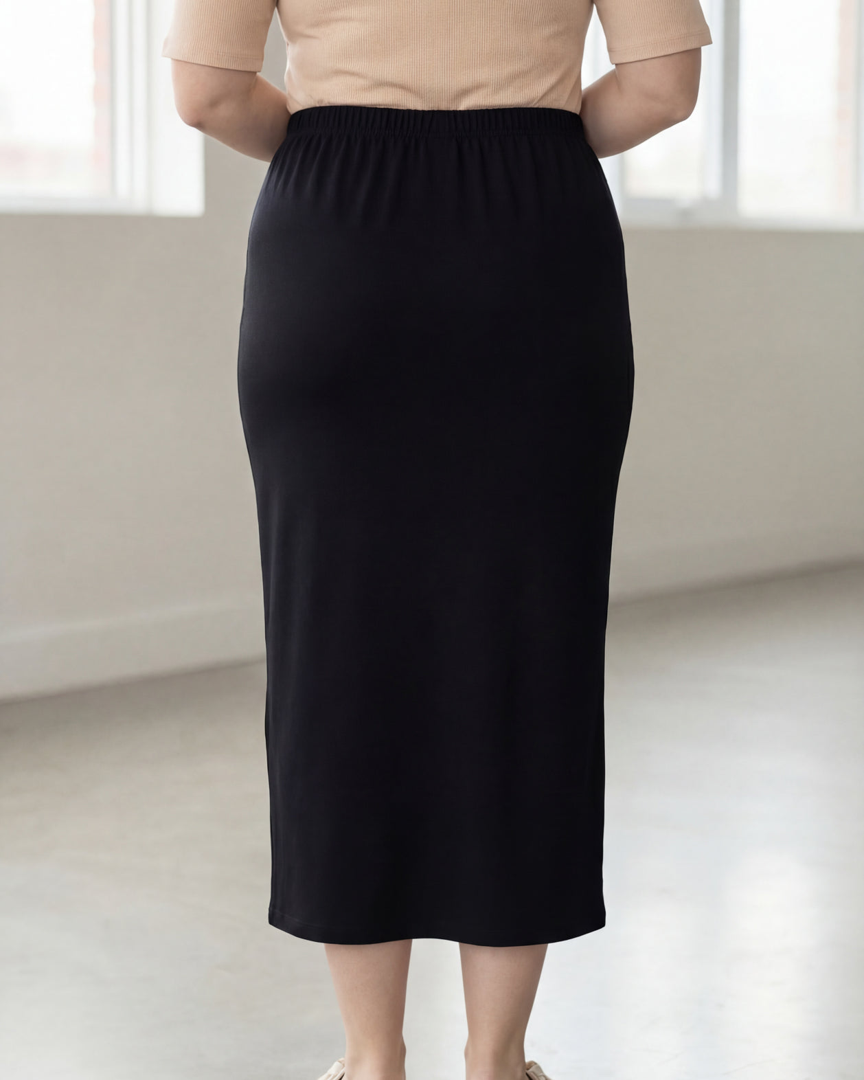Woman wearing a beige shirt & black maxi length pencil skirt standing in a bright room with large windows.