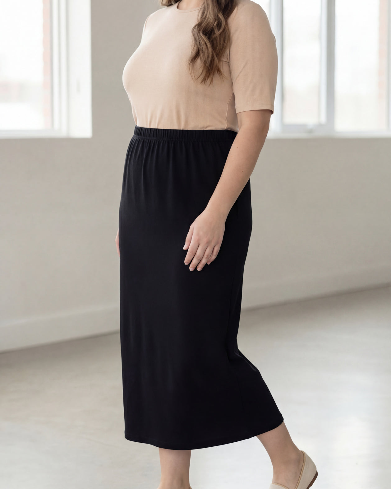 Woman wearing a beige shirt & black maxi length pencil skirt standing in a bright room with large windows.