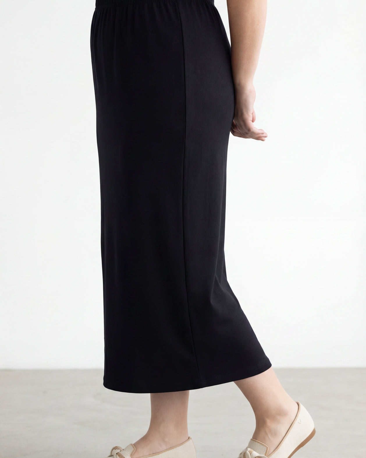 Woman wearing a beige shirt & black maxi length pencil skirt standing in a bright room with large windows.