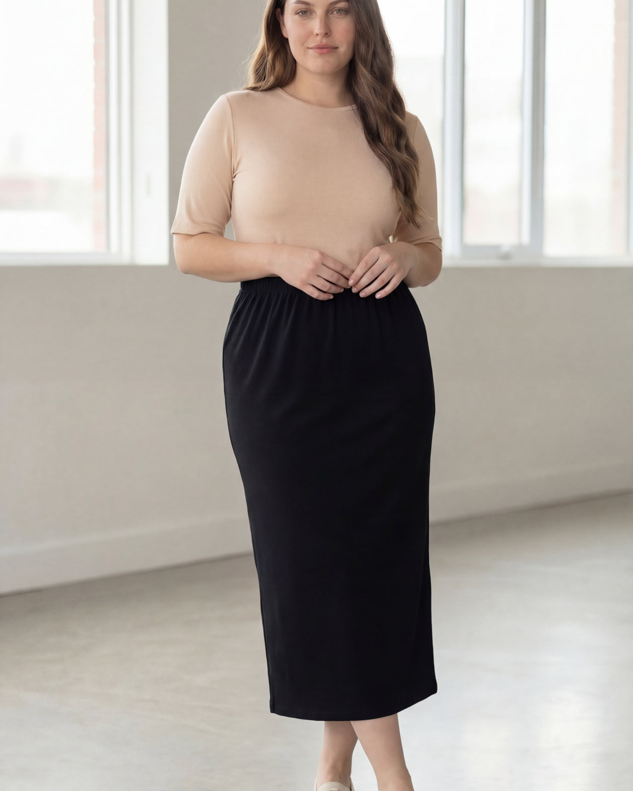 Woman wearing a beige shirt & black maxi length pencil skirt standing in a bright room with large windows.