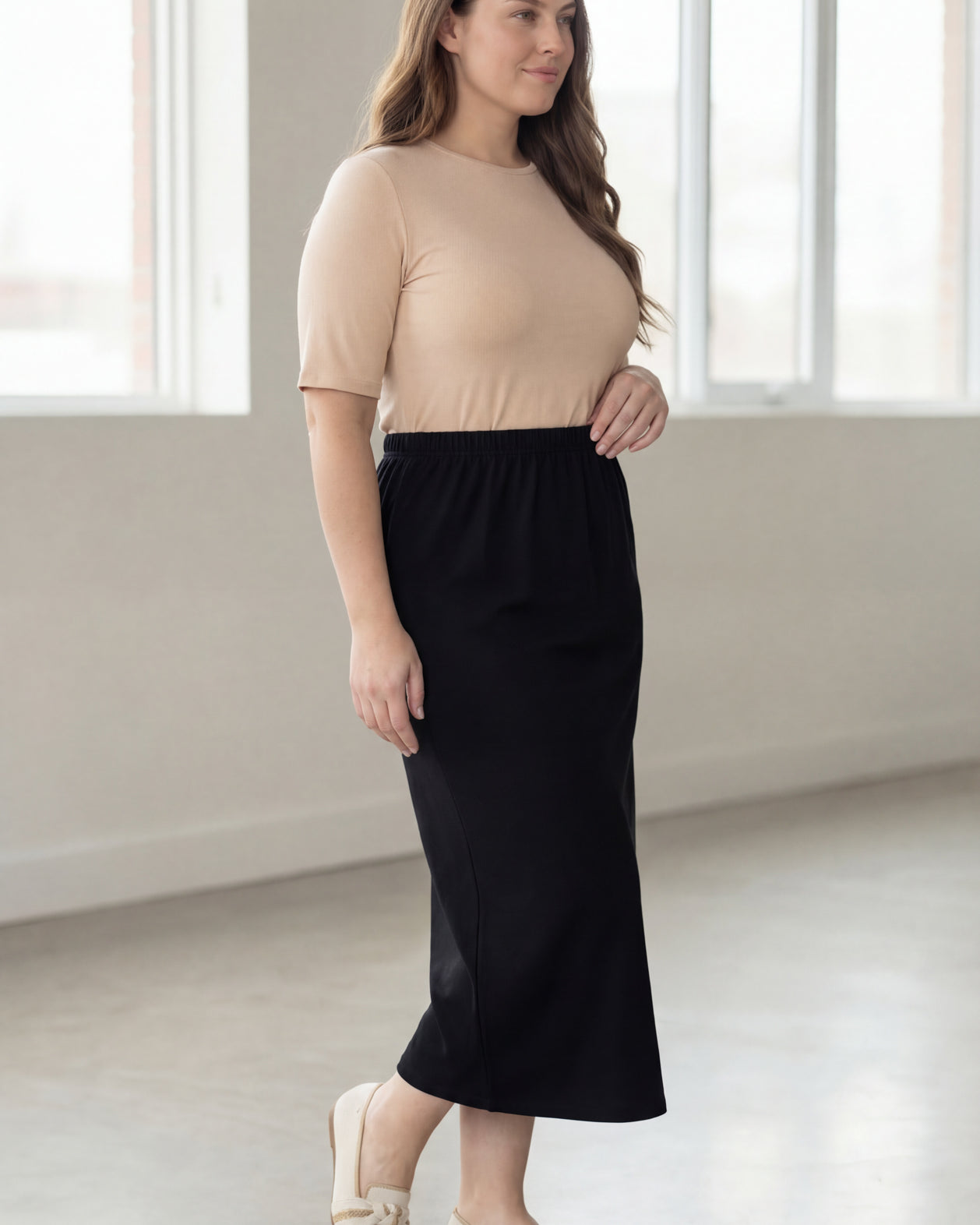 Woman wearing a beige shirt & black maxi length pencil skirt standing in a bright room with large windows.