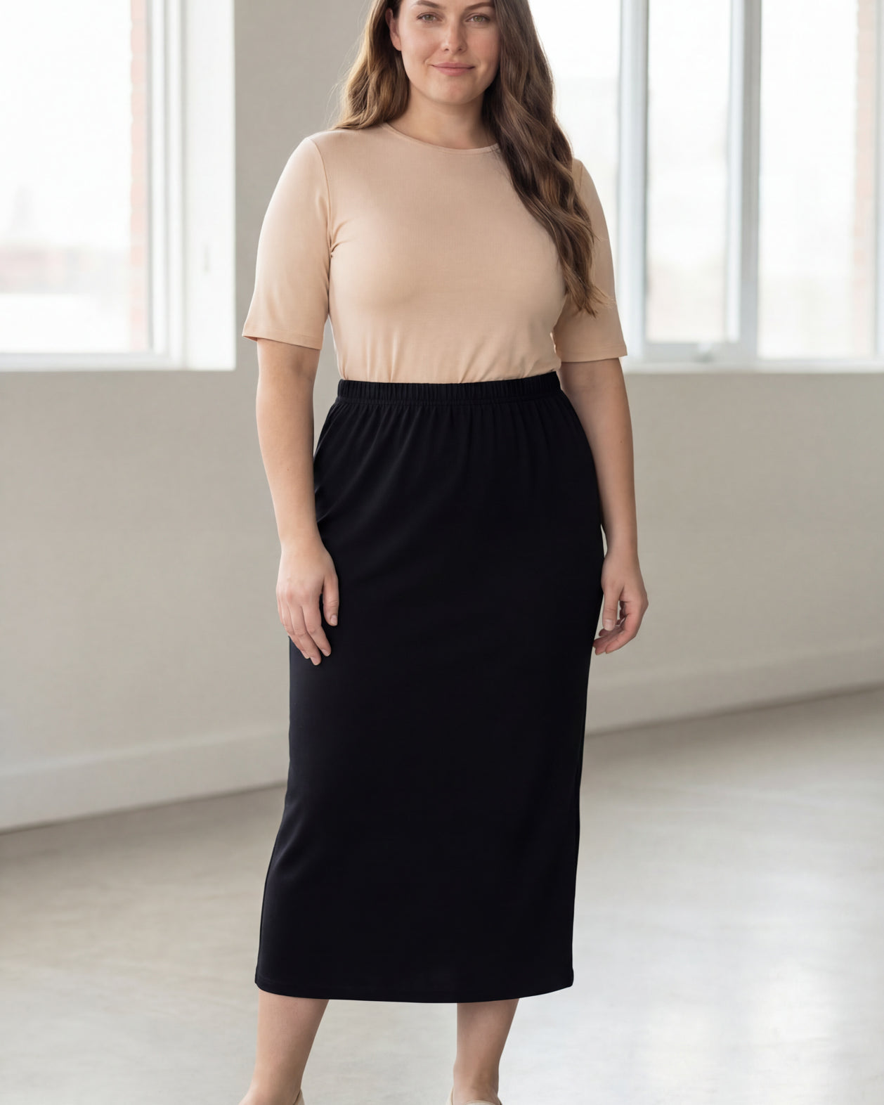 Woman wearing a beige shirt & black maxi length pencil skirt standing in a bright room with large windows.