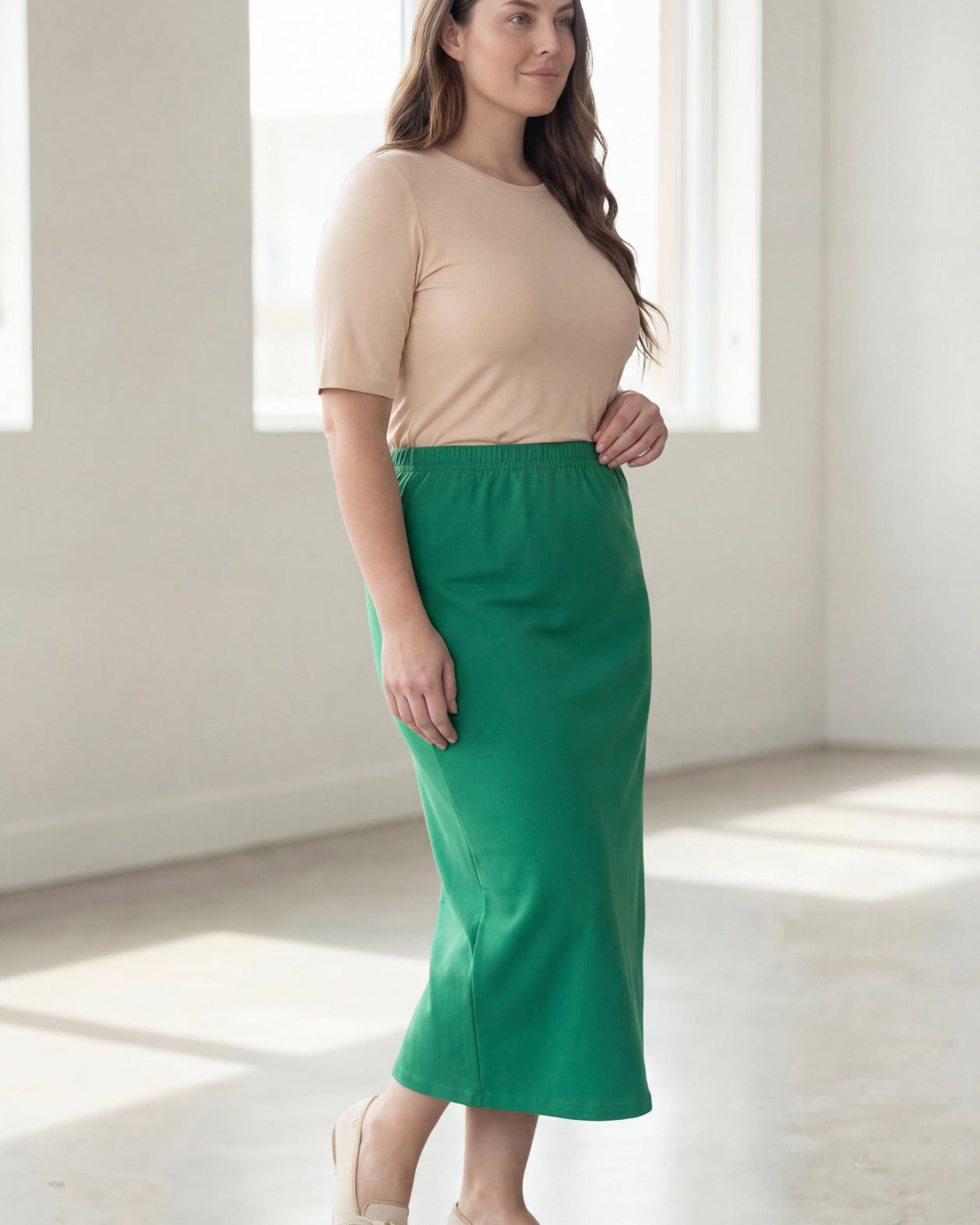 Woman wearing a beige shirt & green maxi length pencil skirt standing in a bright room with large windows.