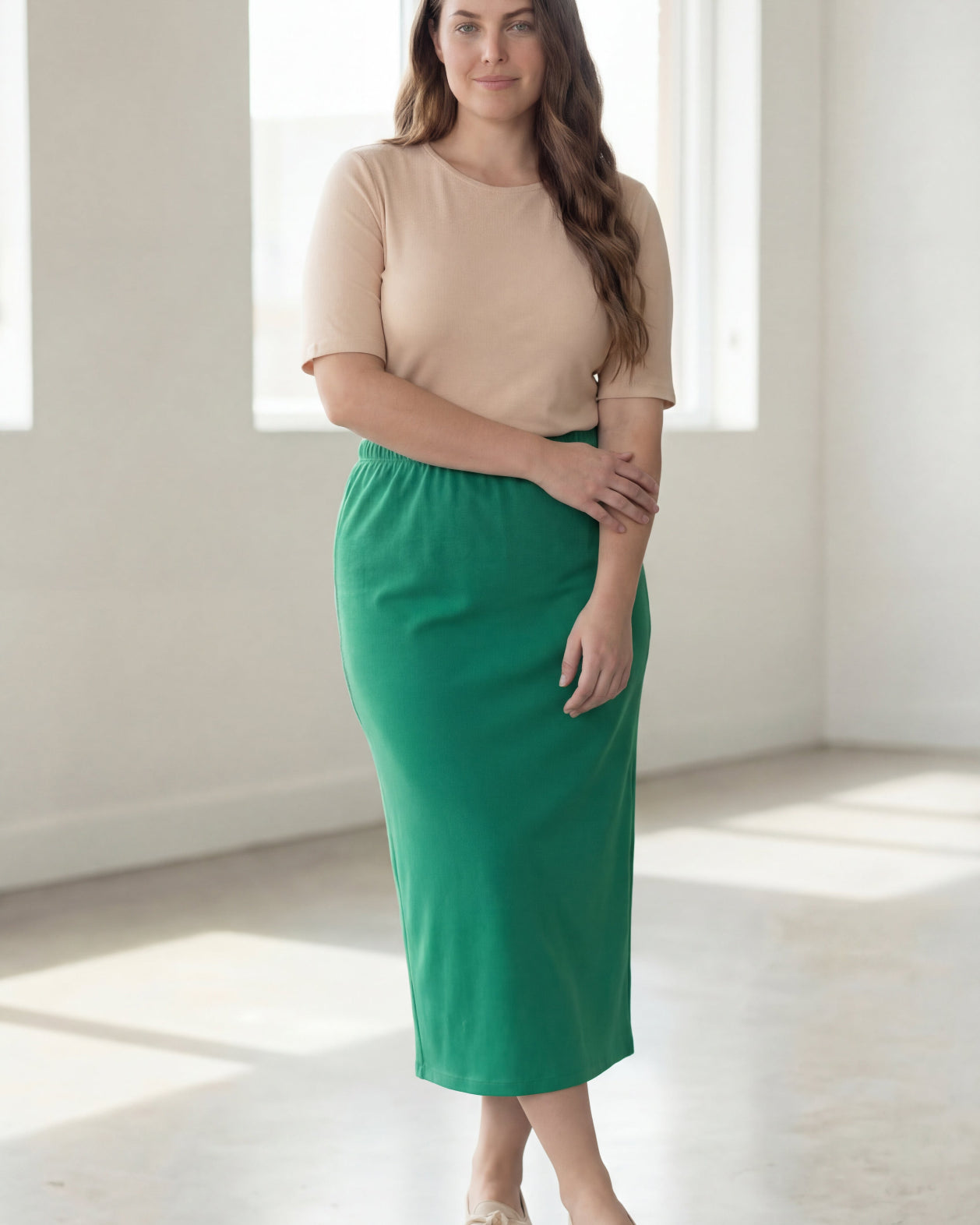 Woman wearing a beige shirt & green maxi length pencil skirt standing in a bright room with large windows.
