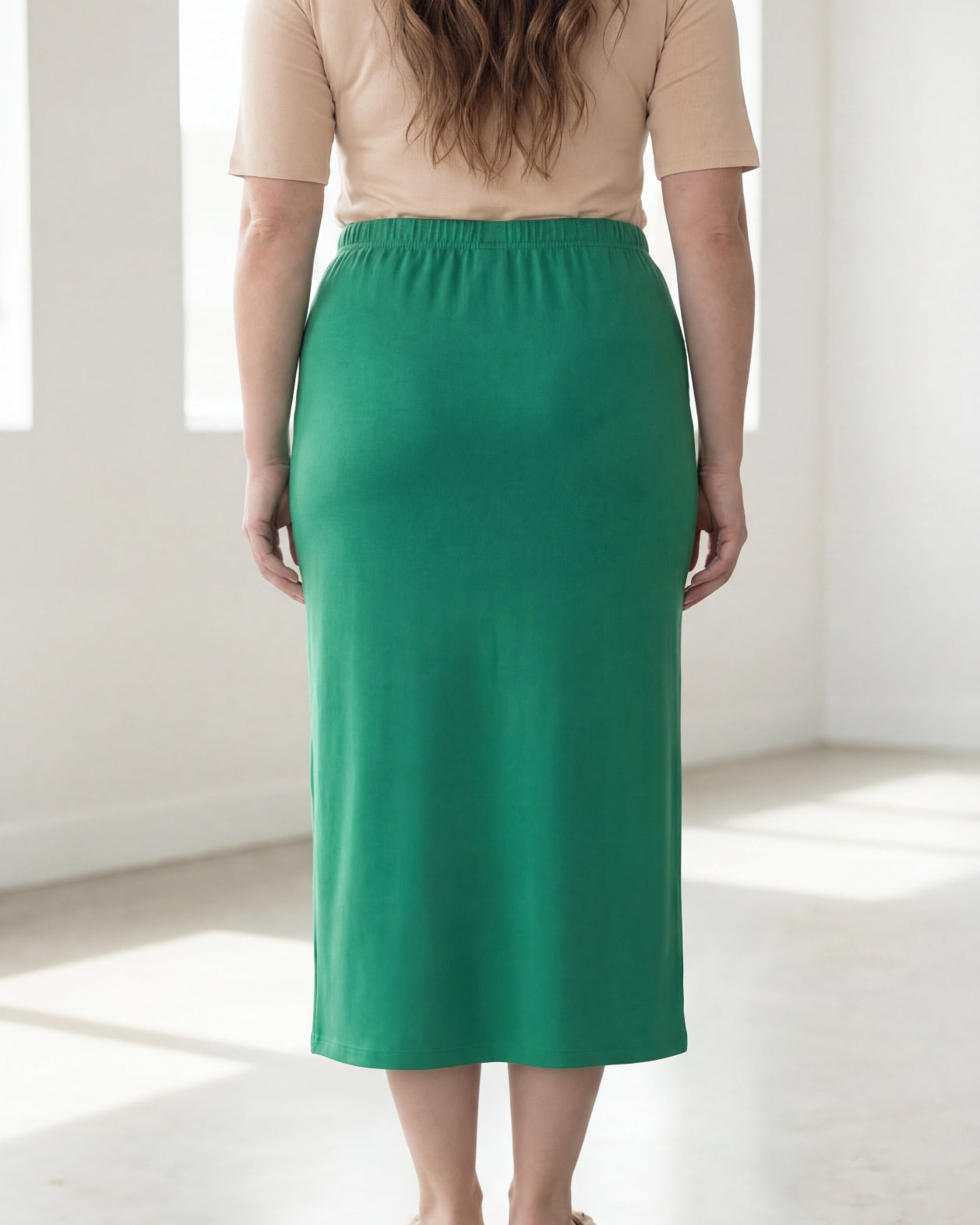 Woman wearing a beige shirt & green maxi length pencil skirt standing in a bright room with large windows.