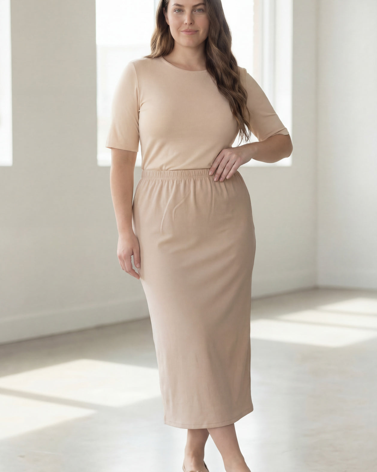Woman wearing a beige shirt & beige maxi length pencil skirt standing in a bright room with large windows.