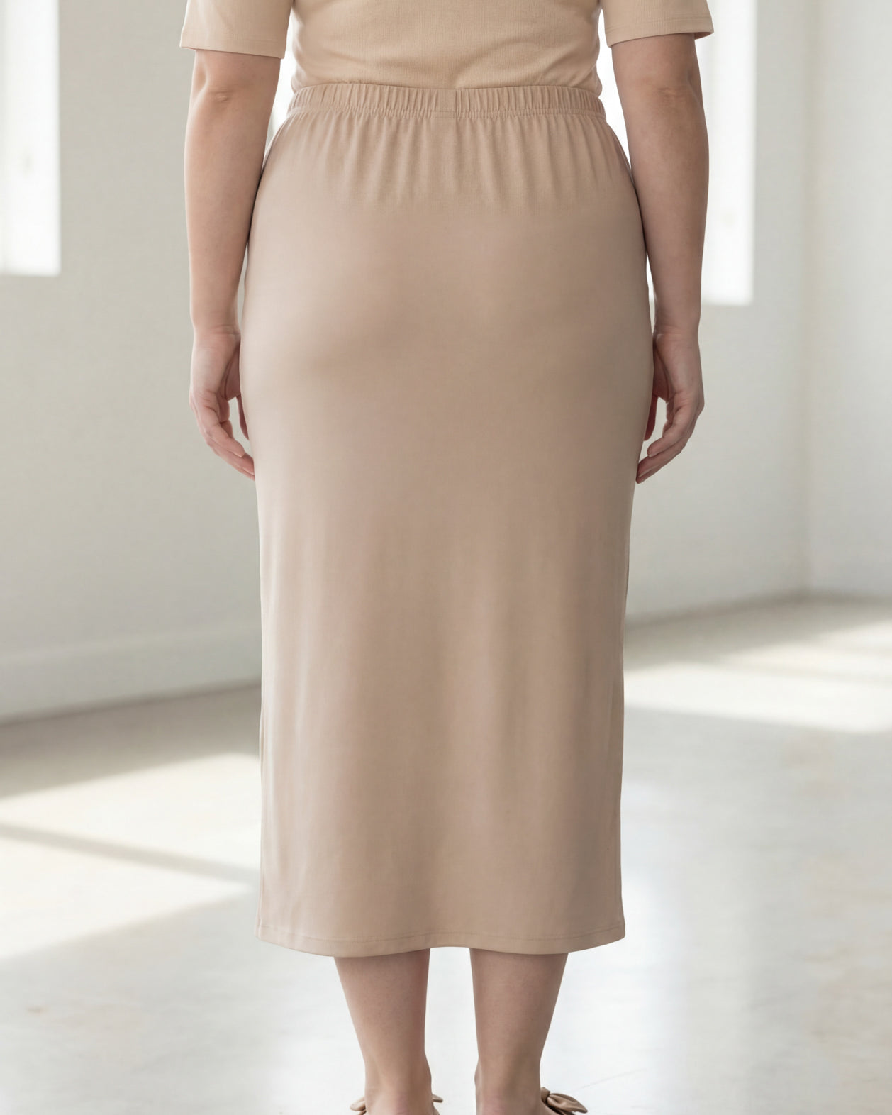 Woman wearing a beige shirt & beige maxi length pencil skirt standing in a bright room with large windows.