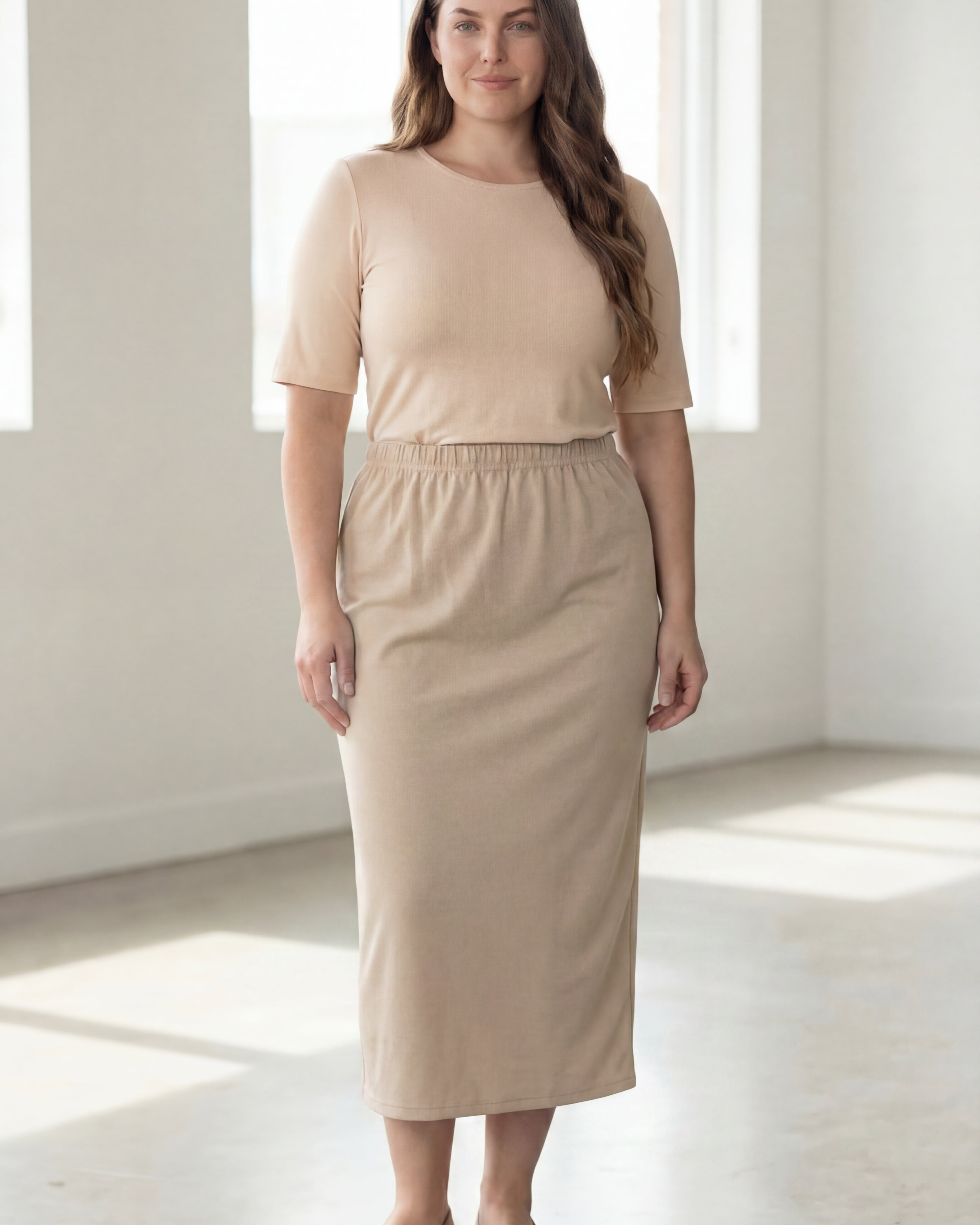Woman wearing a beige shirt & beige maxi length pencil skirt standing in a bright room with large windows.