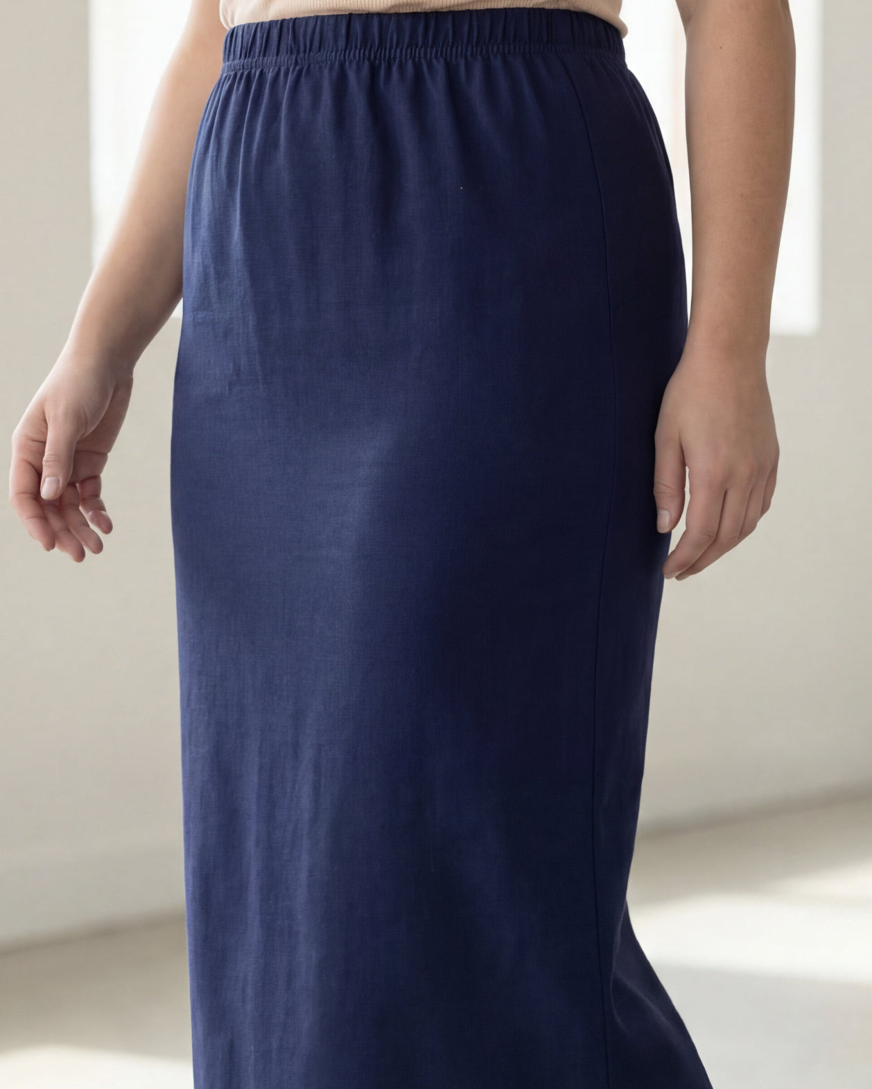 Woman wearing a beige shirt & blue maxi length pencil skirt standing in a bright room with large windows.