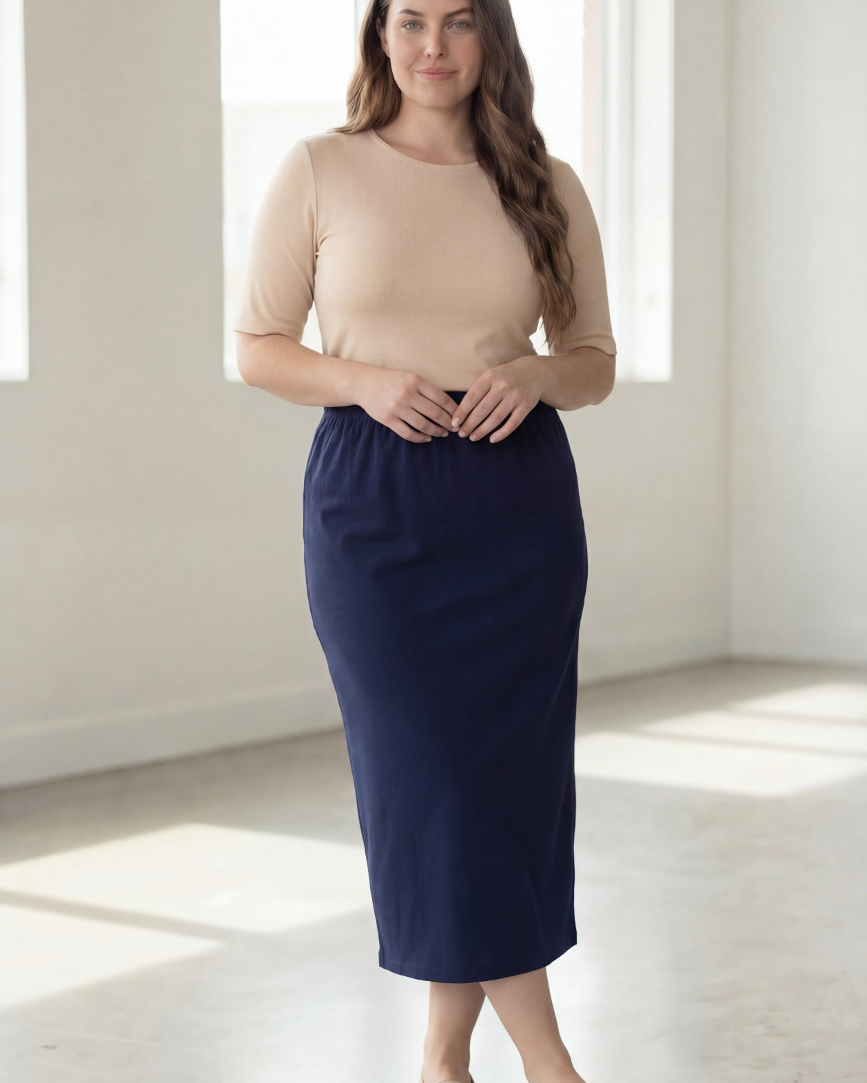 Woman wearing a beige shirt & blue maxi length pencil skirt standing in a bright room with large windows.