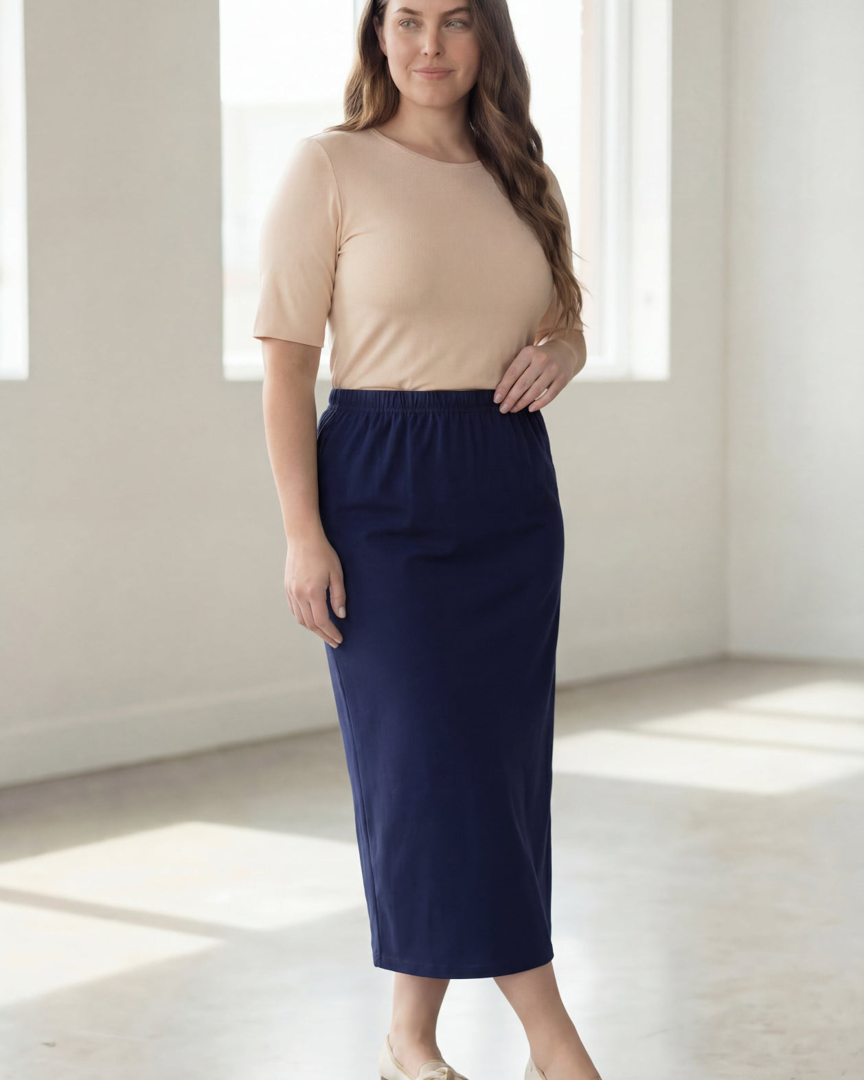 Woman wearing a beige shirt & blue maxi length pencil skirt standing in a bright room with large windows.