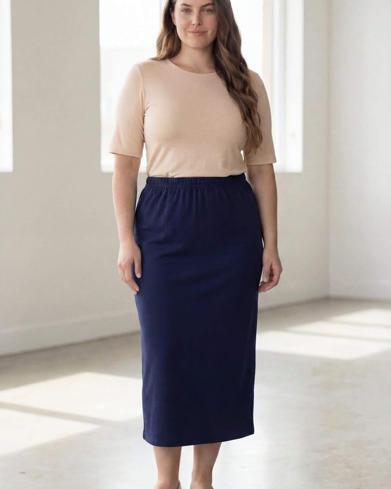Woman wearing a beige shirt & blue maxi length pencil skirt standing in a bright room with large windows.