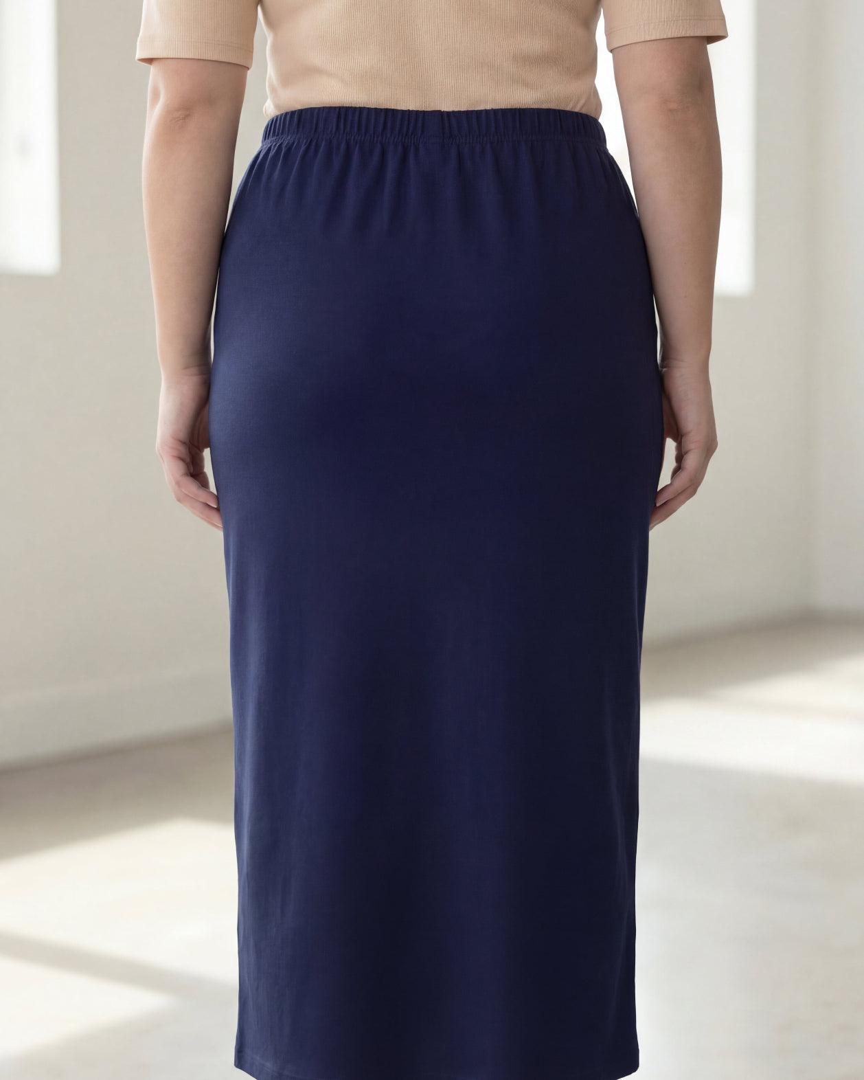 Woman wearing a beige shirt & blue maxi length pencil skirt standing in a bright room with large windows.