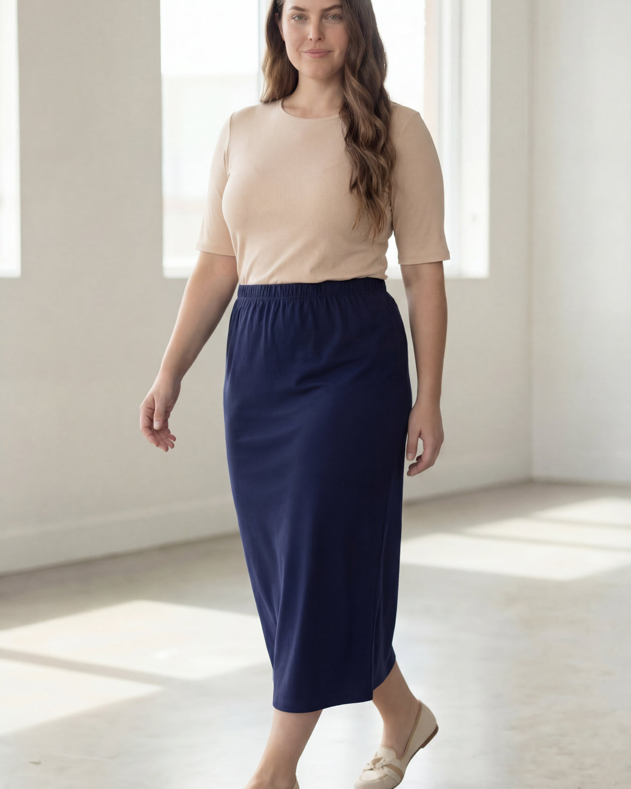 Woman wearing a beige shirt & blue maxi length pencil skirt standing in a bright room with large windows.