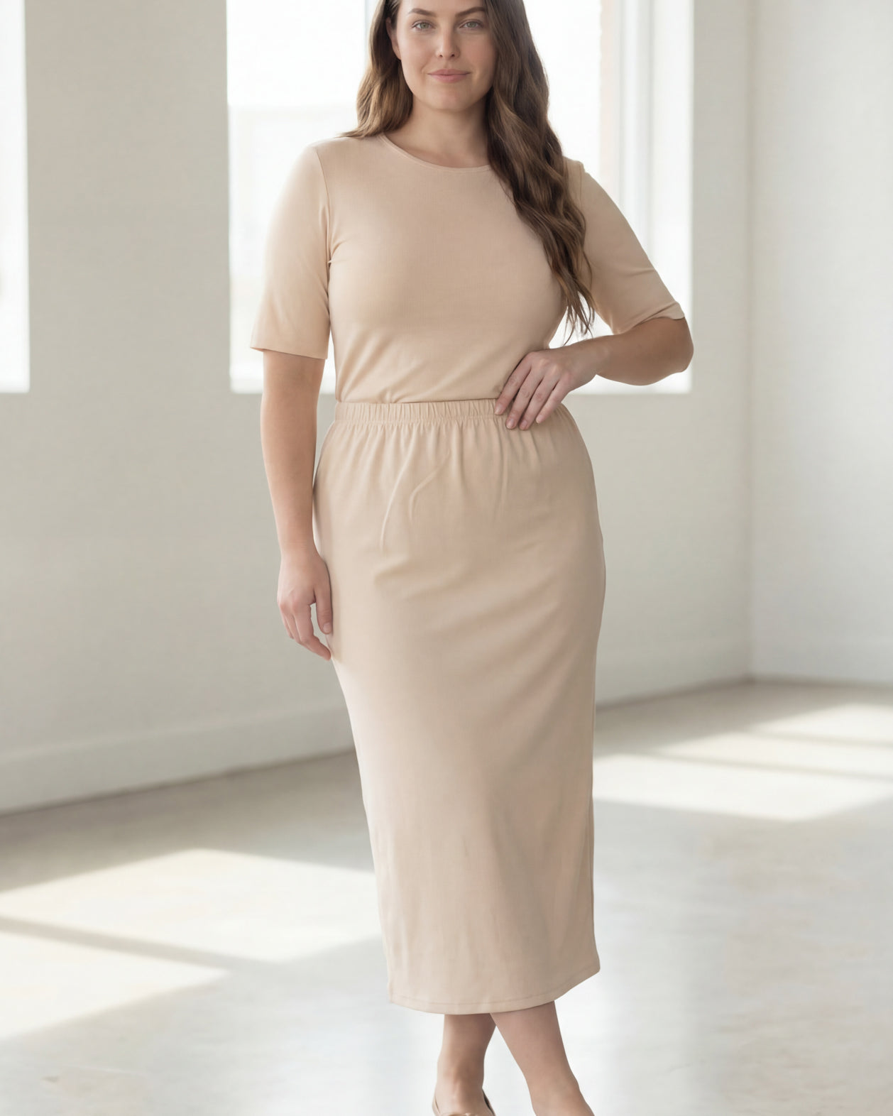 Woman wearing a beige shirt & maxi length pencil skirt standing in a bright room with large windows.