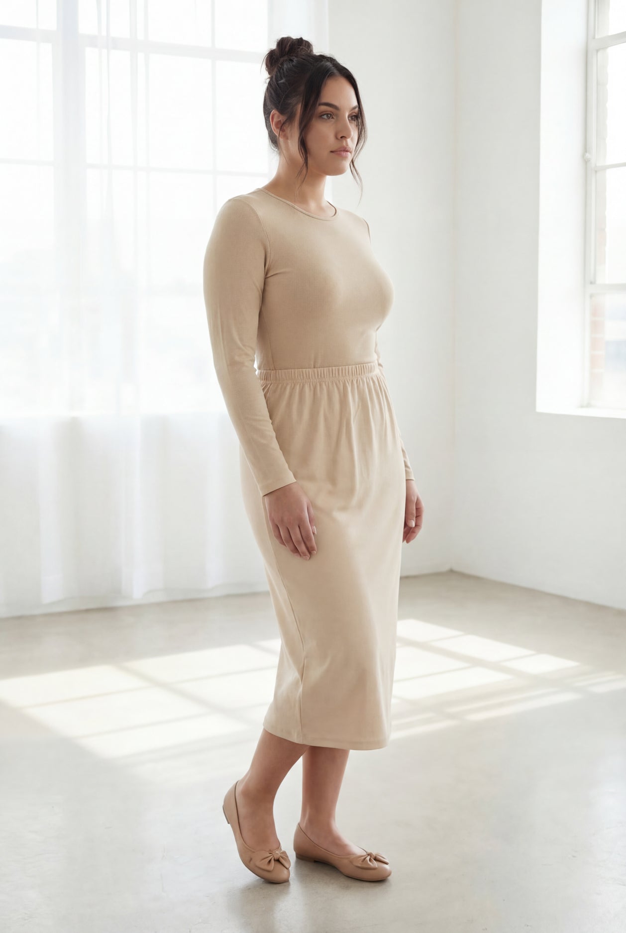 Woman wearing a beige shirt & skirt standing in a bright, minimalistic room.