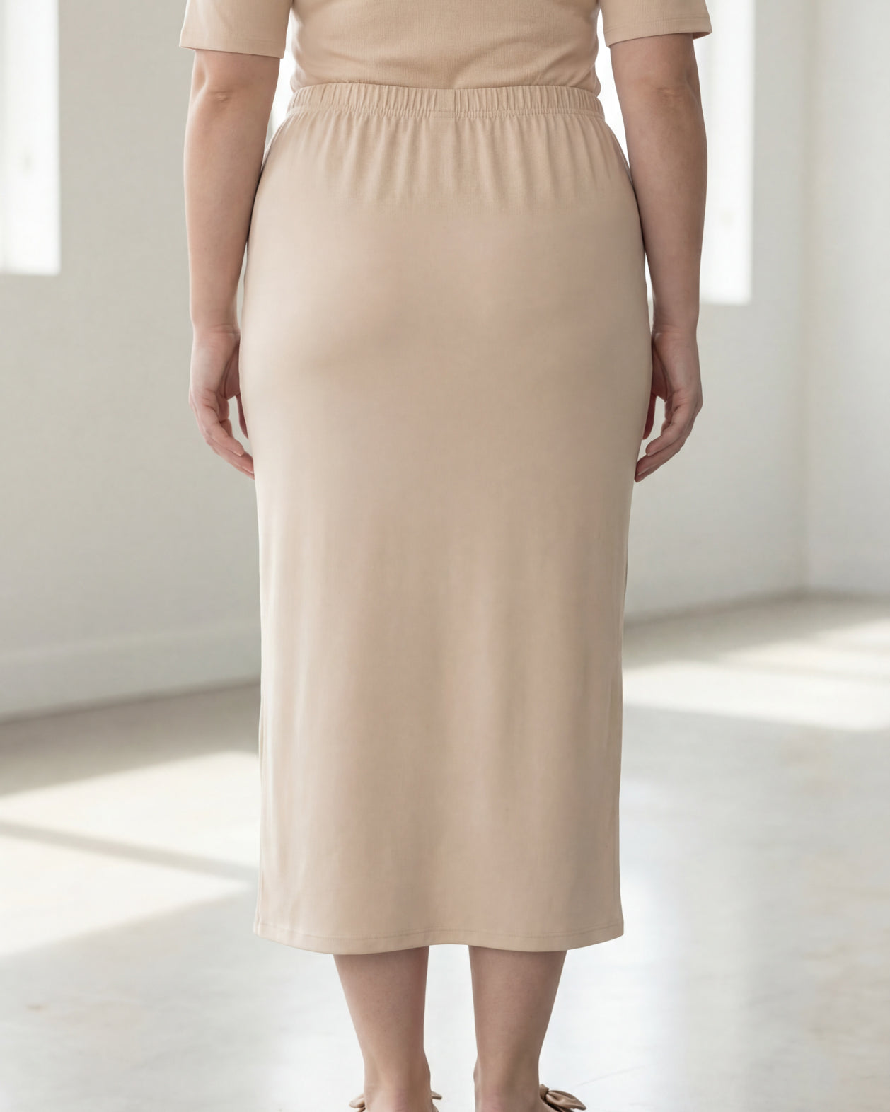 Woman wearing a beige shirt & maxi length pencil skirt standing in a bright room with large windows.
