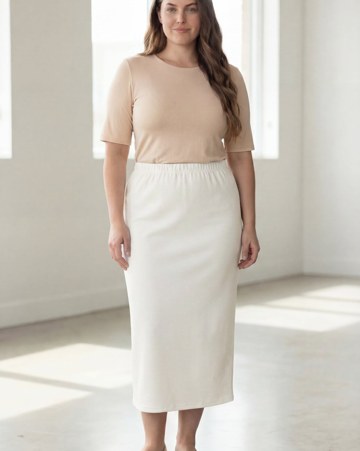Woman wearing a beige shirt & off white maxi length pencil skirt standing in a bright room with large windows.