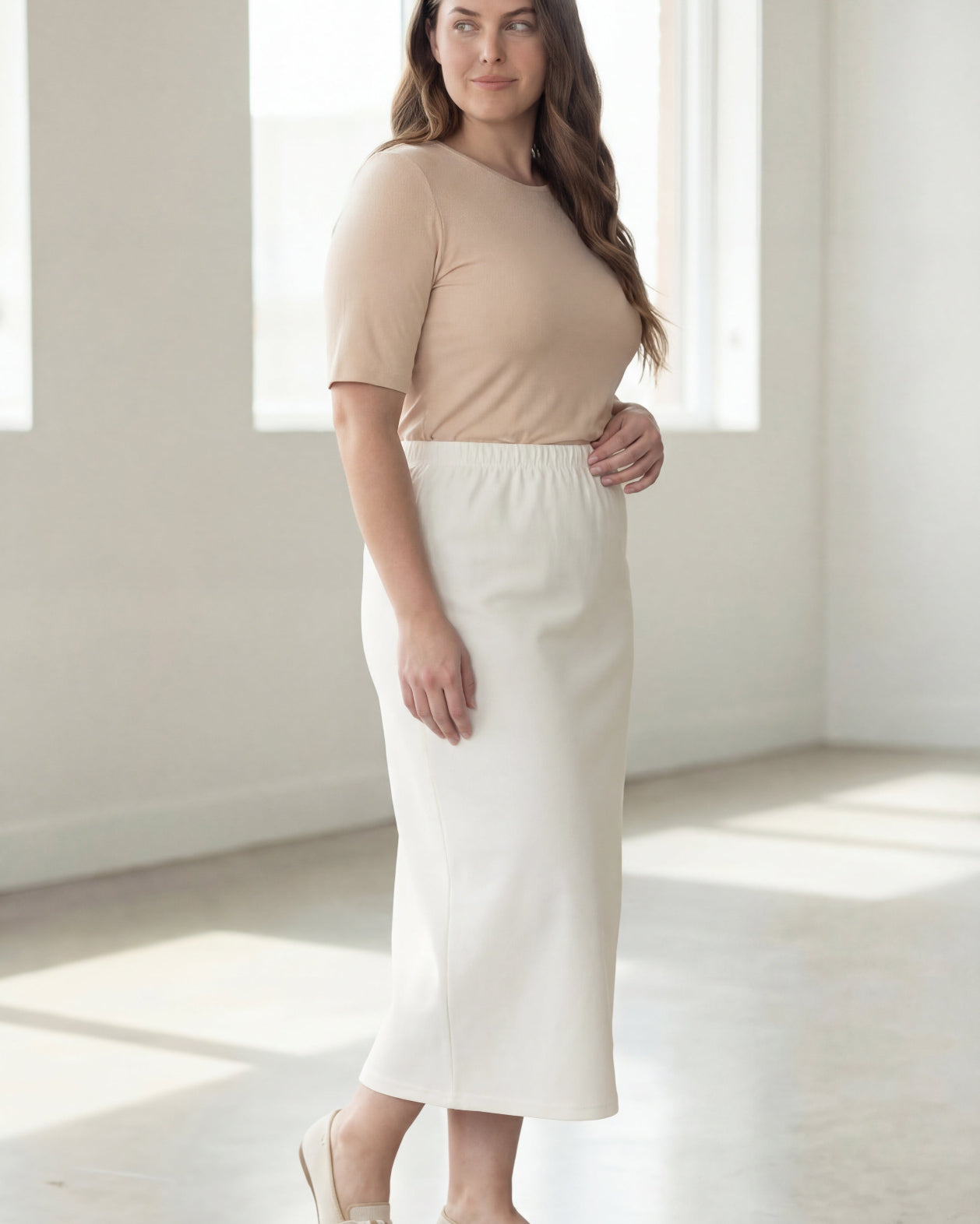 Woman wearing a beige shirt & off white maxi length pencil skirt standing in a bright room with large windows.