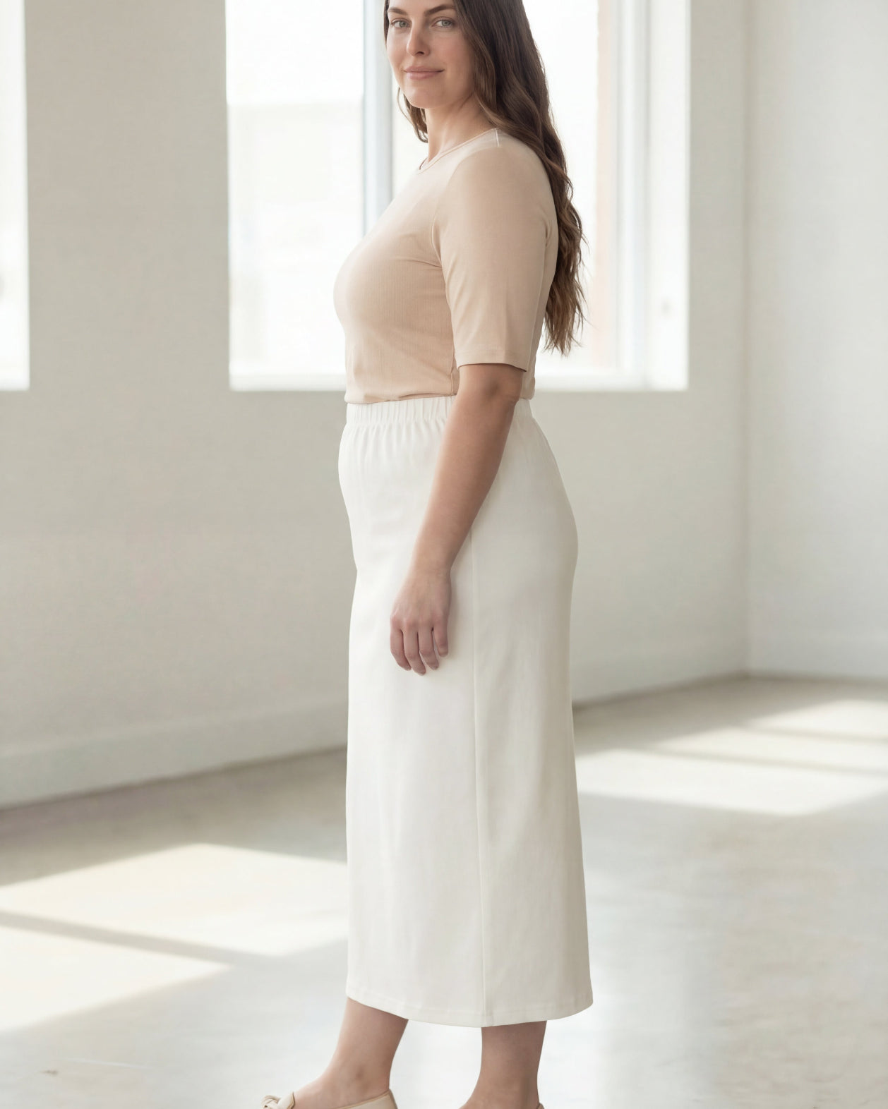 Woman wearing a beige shirt & off white maxi length pencil skirt standing in a bright room with large windows.
