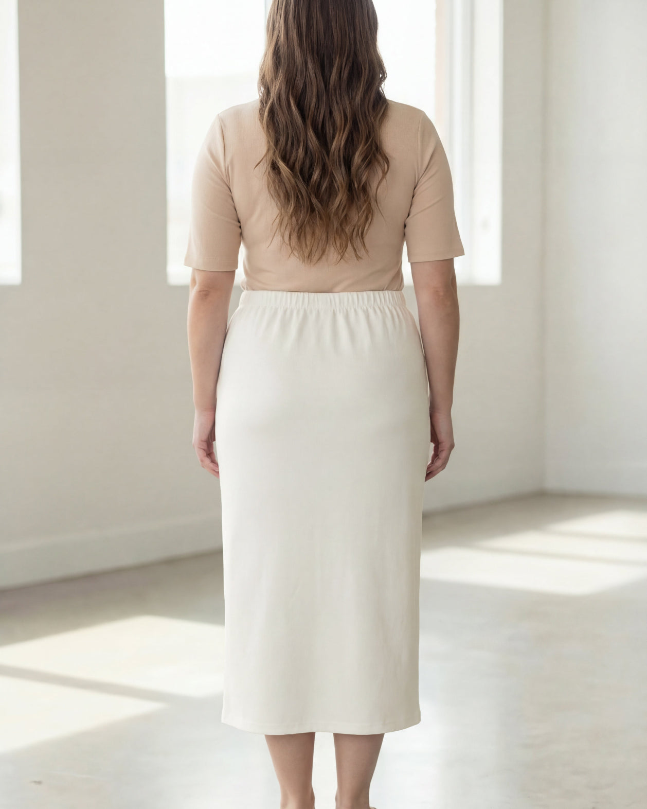 Woman wearing a beige shirt & off white maxi length pencil skirt standing in a bright room with large windows.
