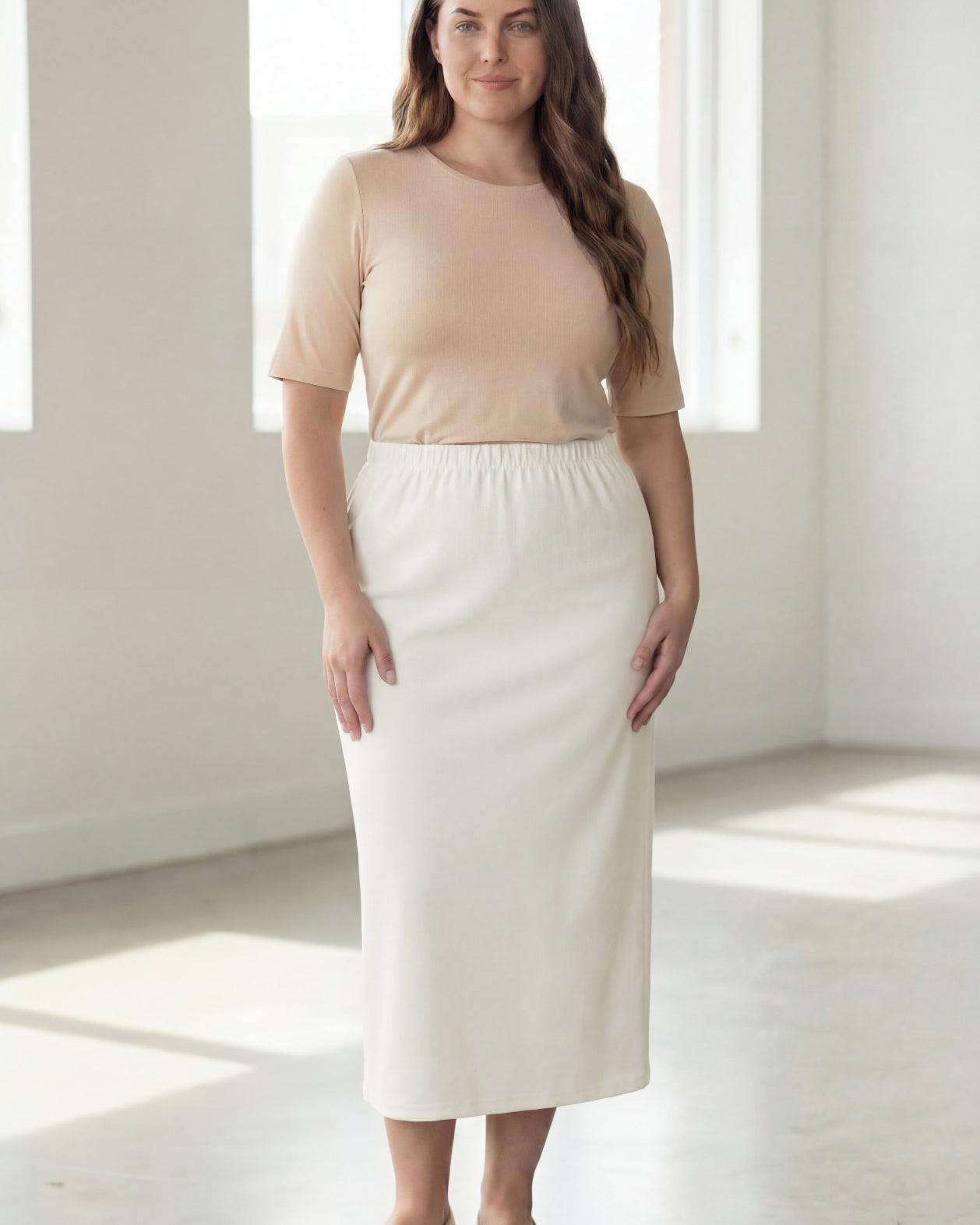 Woman wearing a beige shirt & off white maxi length pencil skirt standing in a bright room with large windows.