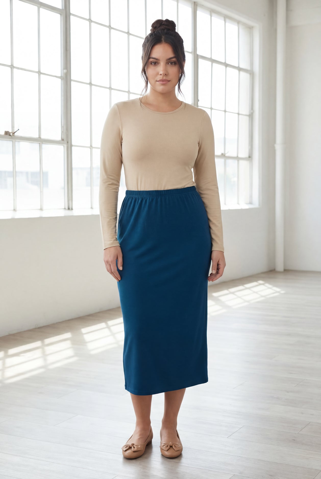 Woman wearing a beige top and blue skirt in a bright room with large windows.