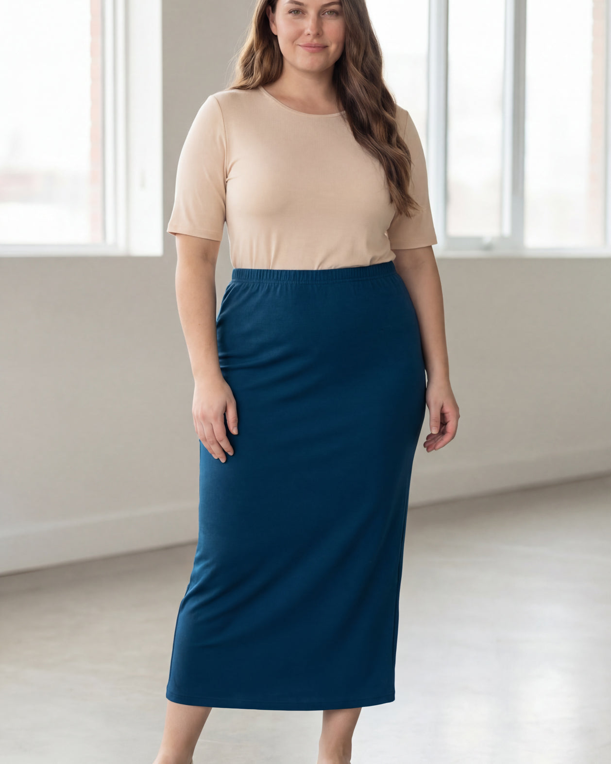 Woman wearing a beige shirt & blue maxi length pencil skirt standing in a bright room with large windows.