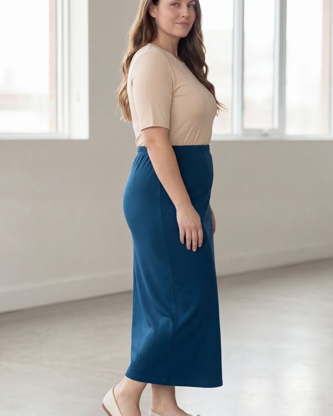 Woman wearing a beige shirt & blue maxi length pencil skirt standing in a bright room with large windows.