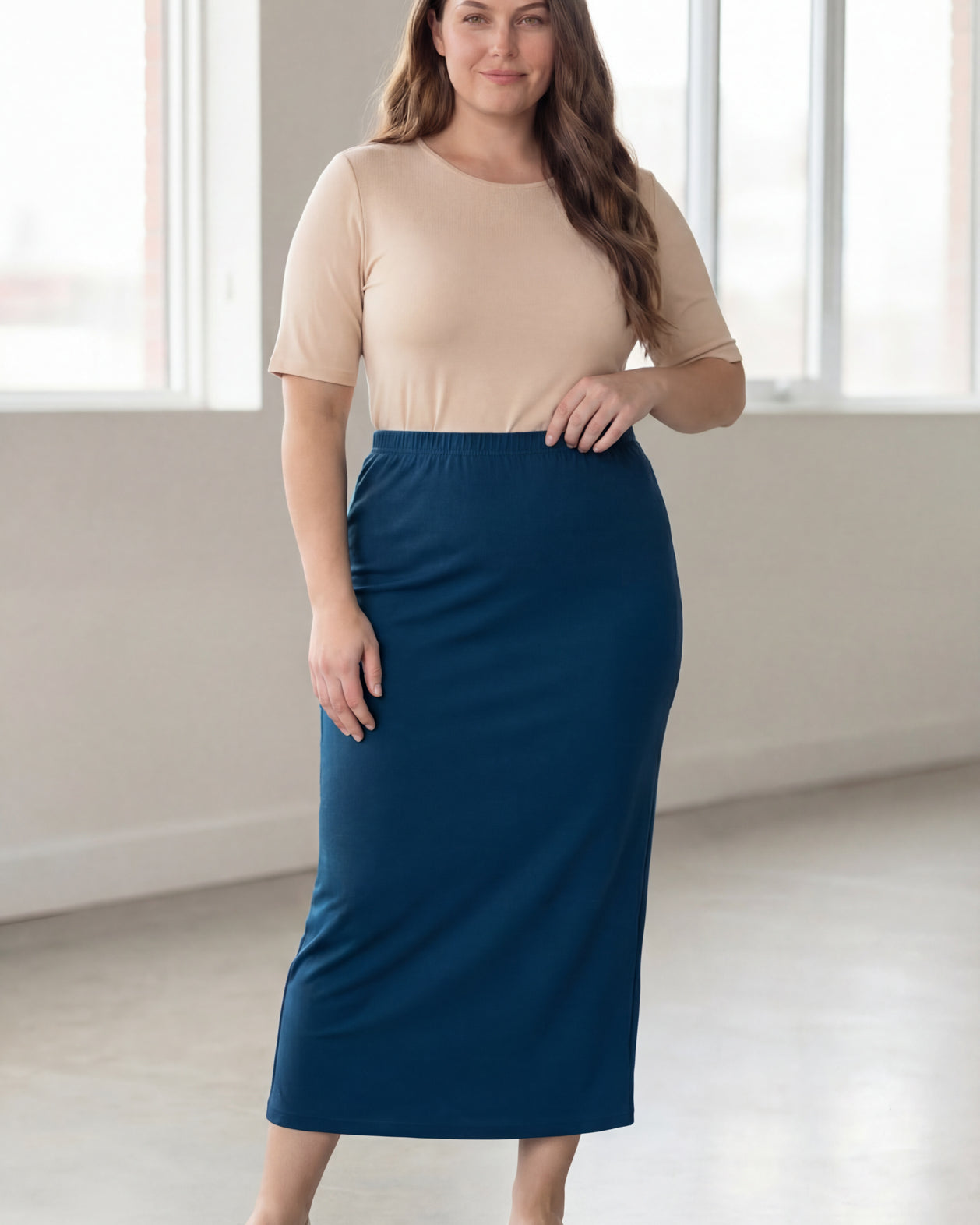 Woman wearing a beige shirt & blue maxi length pencil skirt standing in a bright room with large windows.