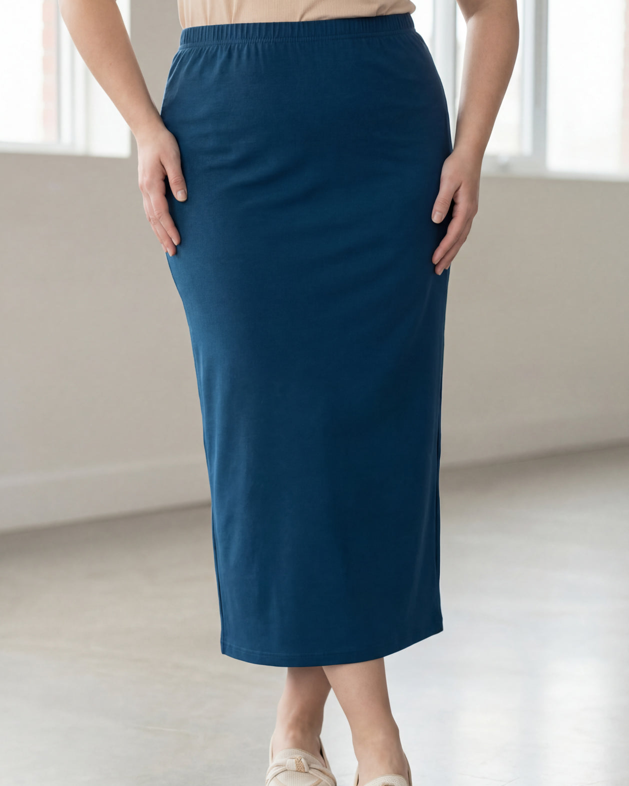 Woman wearing a beige shirt & blue maxi length pencil skirt standing in a bright room with large windows.