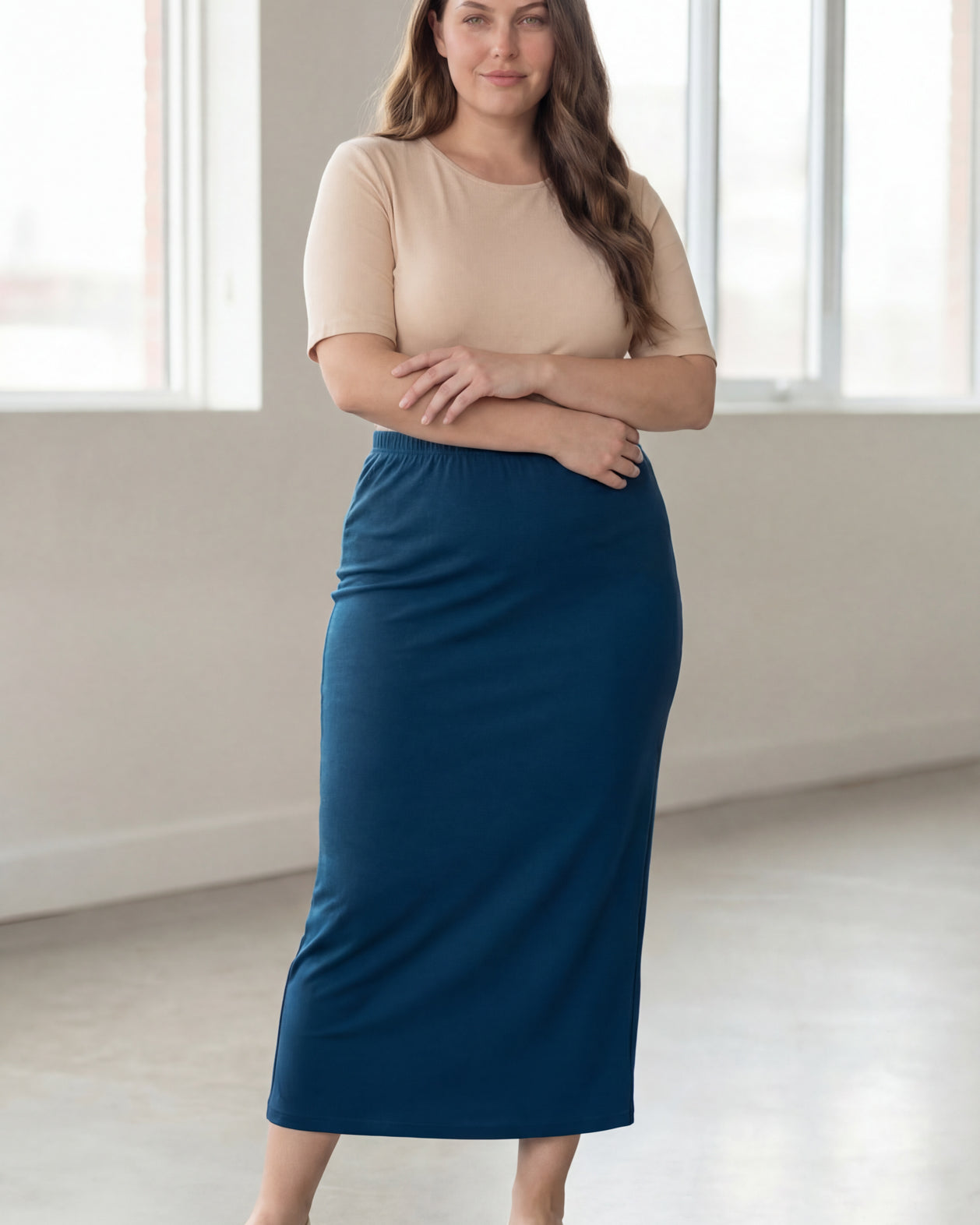 Woman wearing a beige shirt & blue maxi length pencil skirt standing in a bright room with large windows.