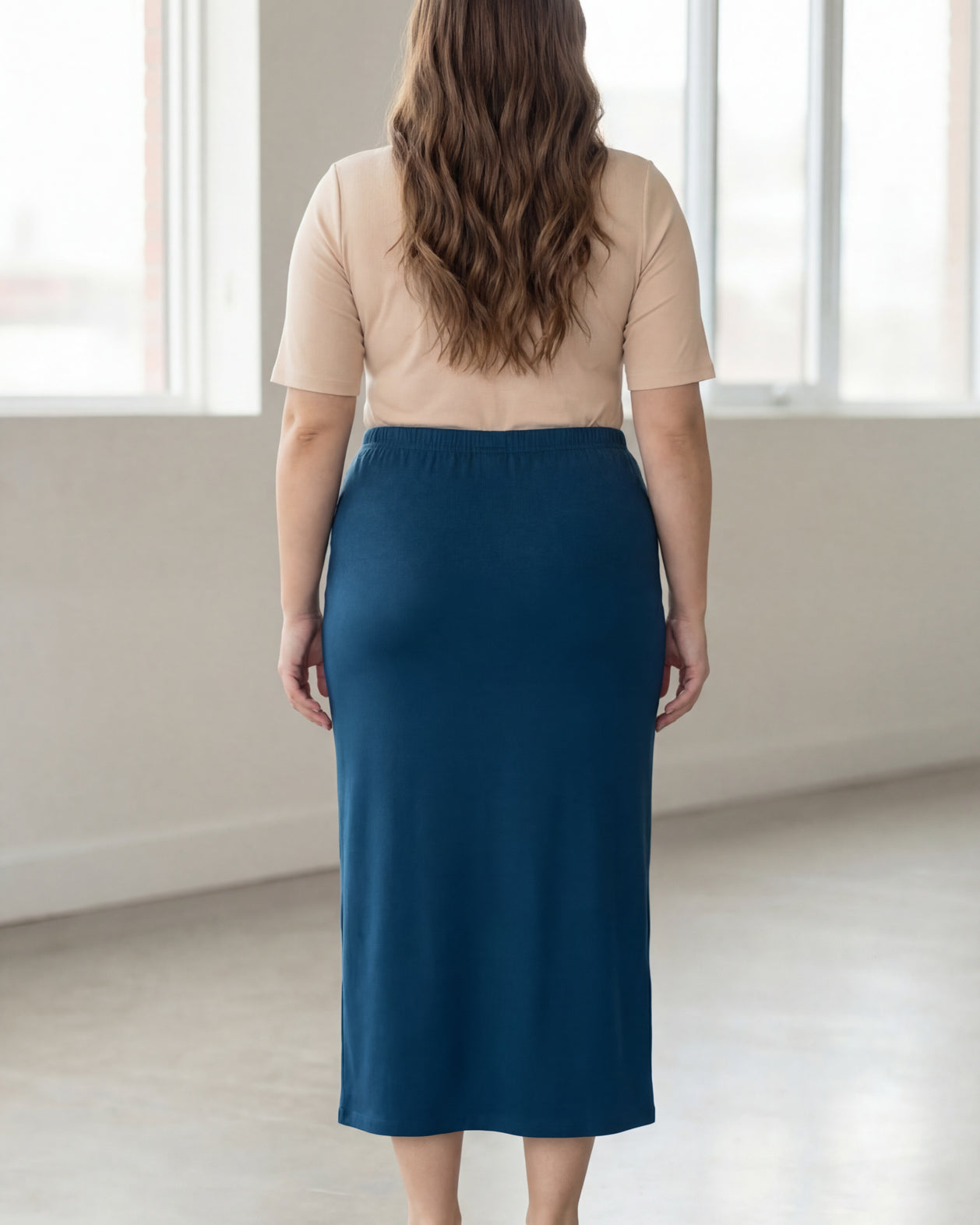 Woman wearing a beige shirt & blue maxi length pencil skirt standing in a bright room with large windows.