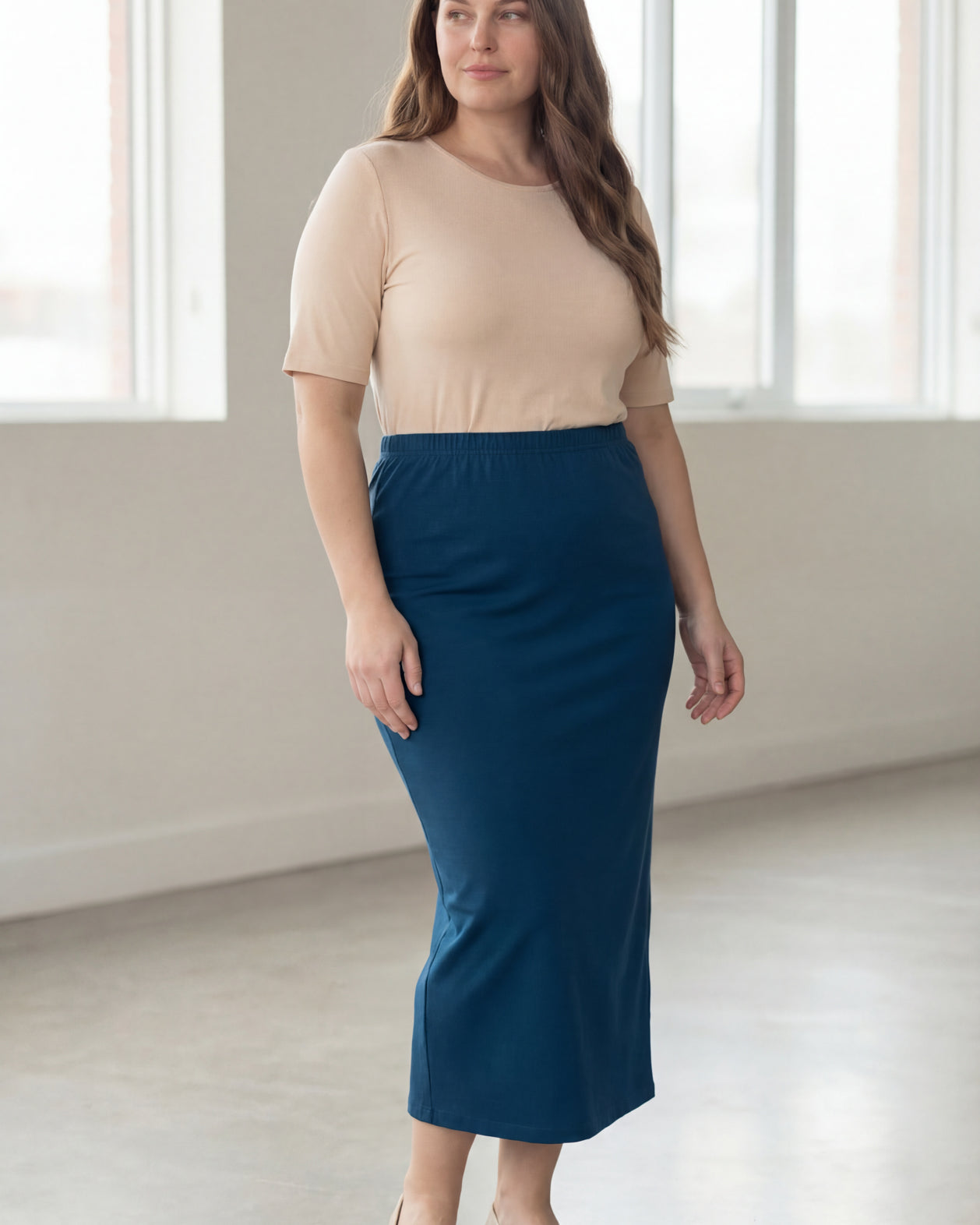 Woman wearing a beige shirt & blue maxi length pencil skirt standing in a bright room with large windows.