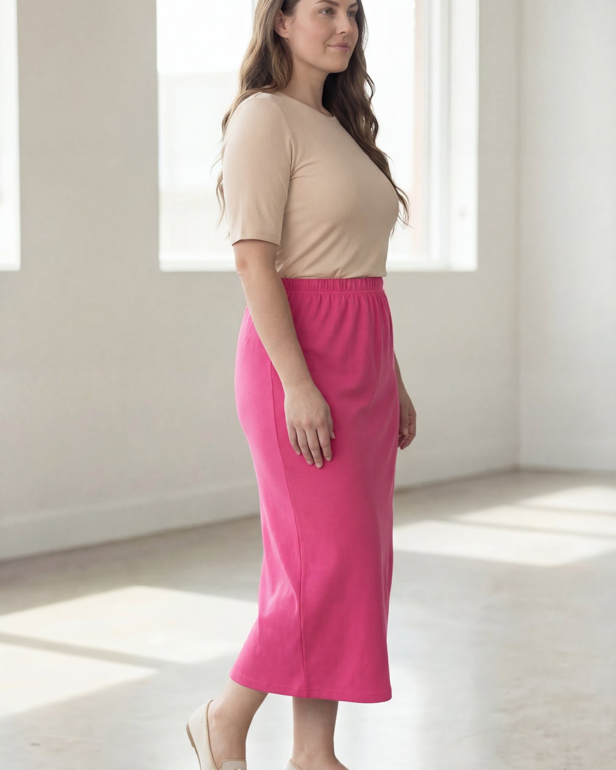 Woman wearing a beige shirt & pink maxi length pencil skirt standing in a bright room with large windows.