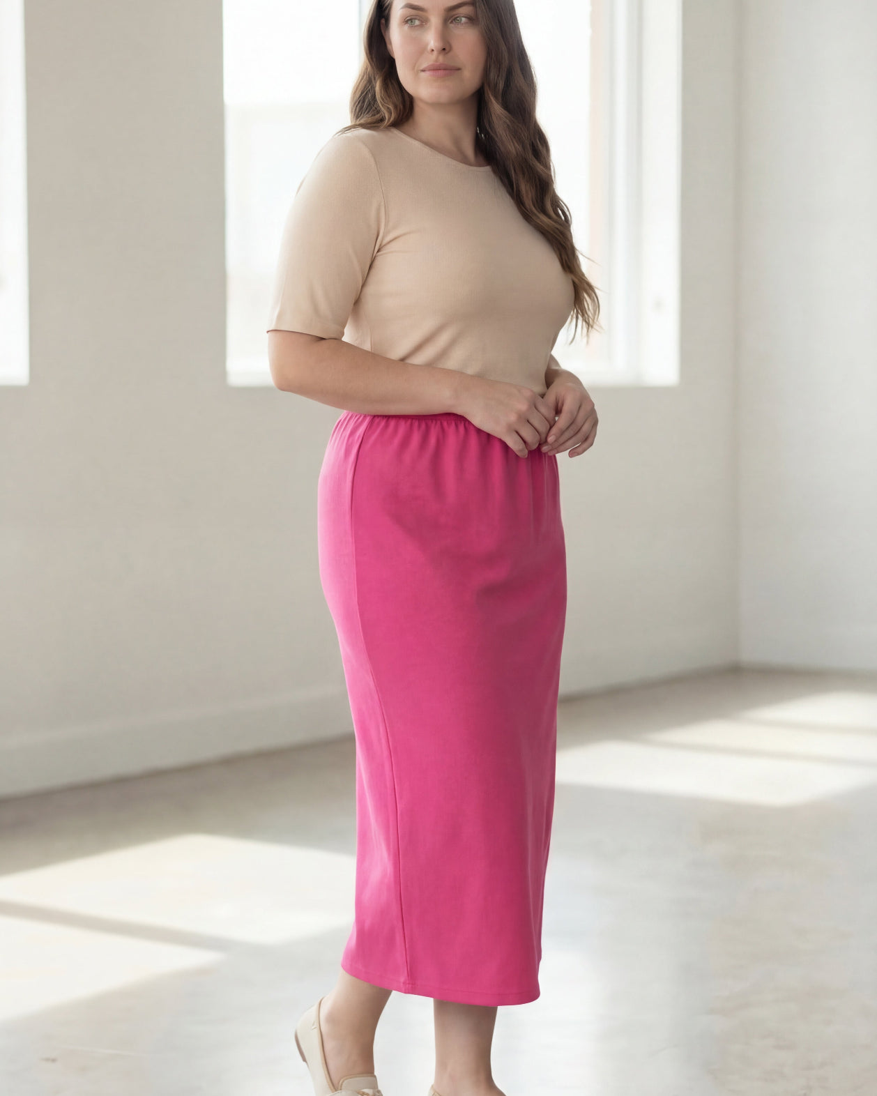 Woman wearing a beige shirt & pink maxi length pencil skirt standing in a bright room with large windows.