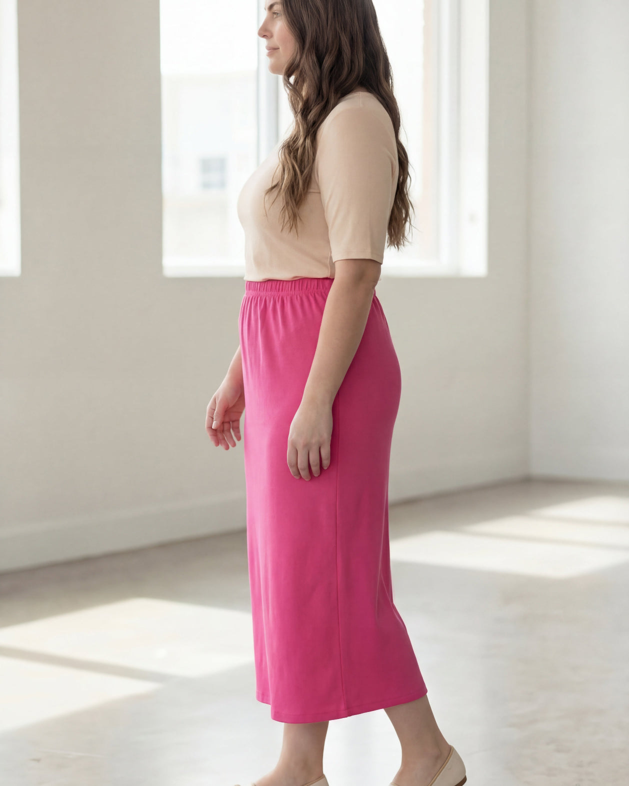 Woman wearing a beige shirt & pink maxi length pencil skirt standing in a bright room with large windows.