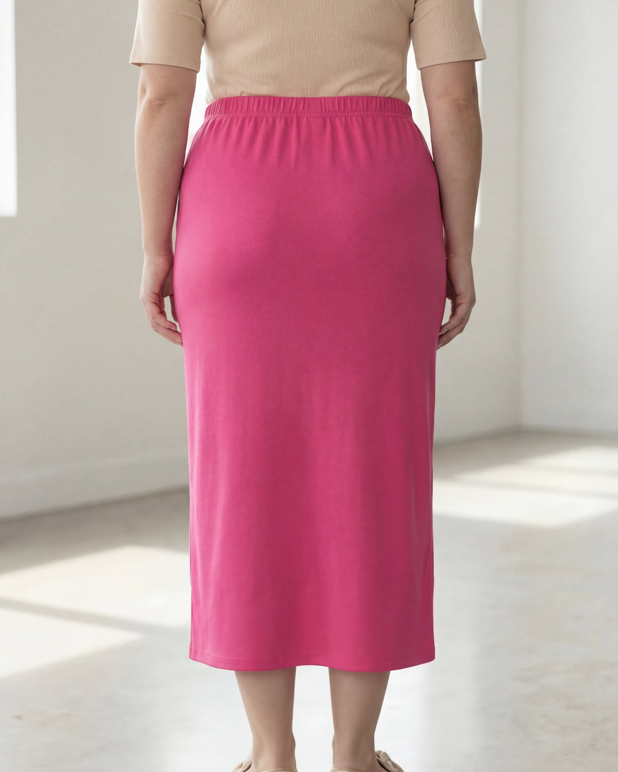 Woman wearing a beige shirt & pink maxi length pencil skirt standing in a bright room with large windows.