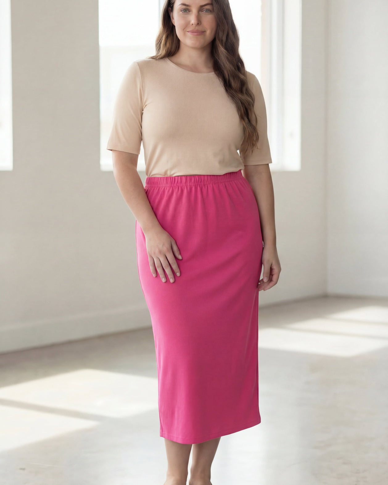 Woman wearing a beige shirt & pink maxi length pencil skirt standing in a bright room with large windows.
