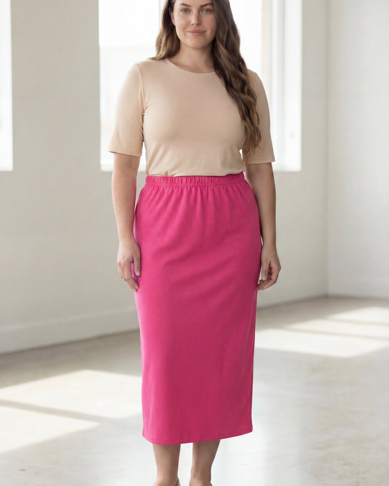 Woman wearing a beige shirt & pink maxi length pencil skirt standing in a bright room with large windows.