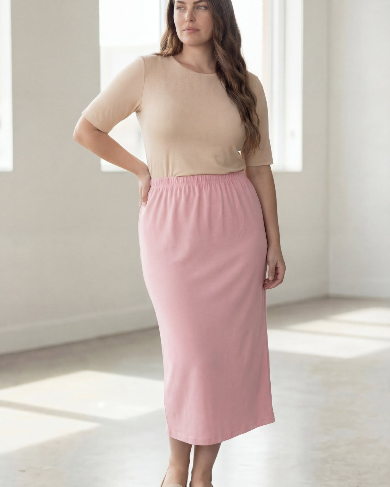 Woman wearing a beige shirt & pink maxi length pencil skirt standing in a bright room with large windows.