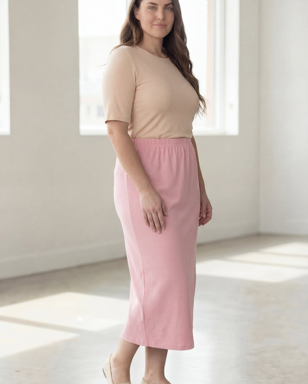 Woman wearing a beige shirt & pink maxi length pencil skirt standing in a bright room with large windows.