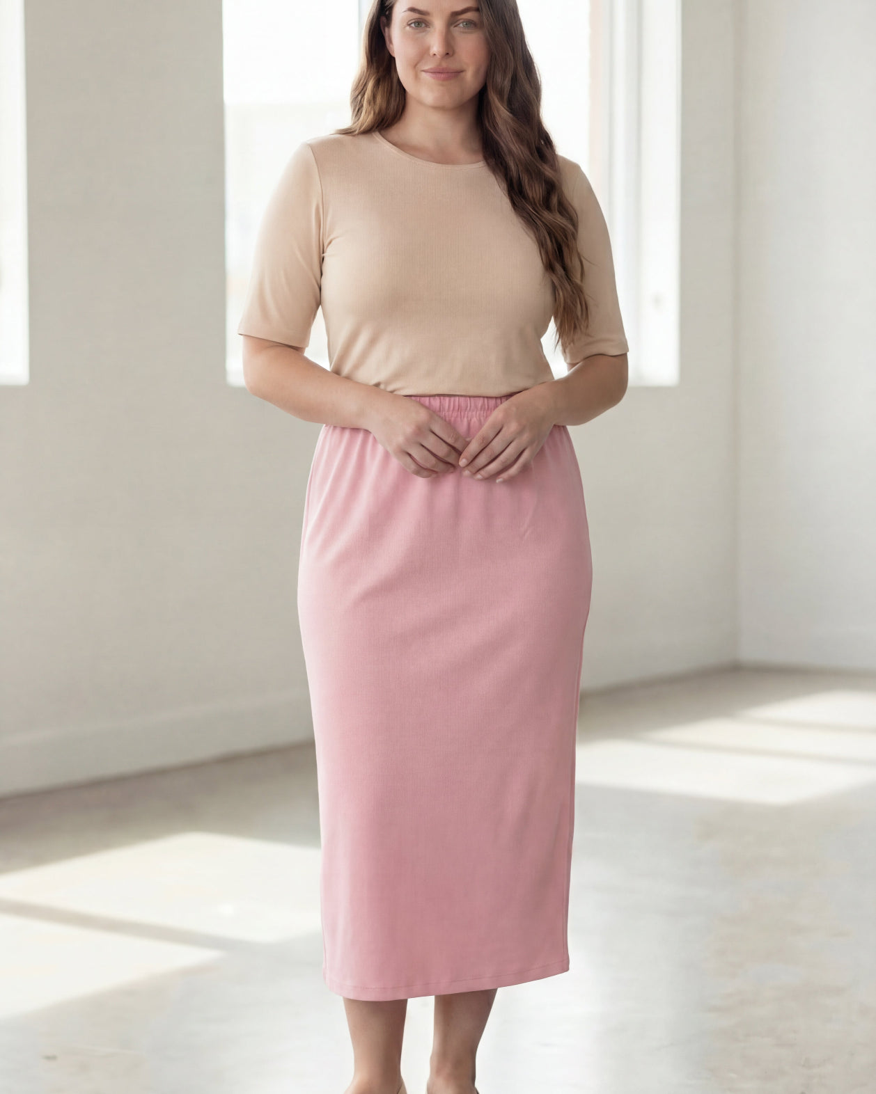 Woman wearing a beige shirt & pink maxi length pencil skirt standing in a bright room with large windows.
