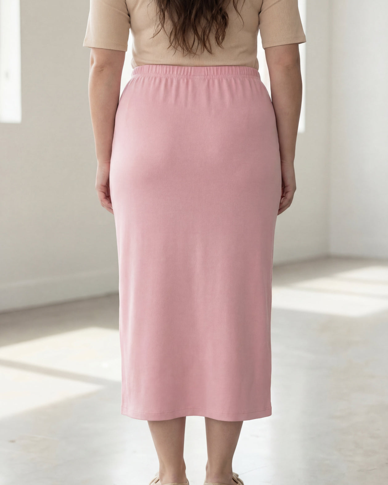 Woman wearing a beige shirt & pink maxi length pencil skirt standing in a bright room with large windows.