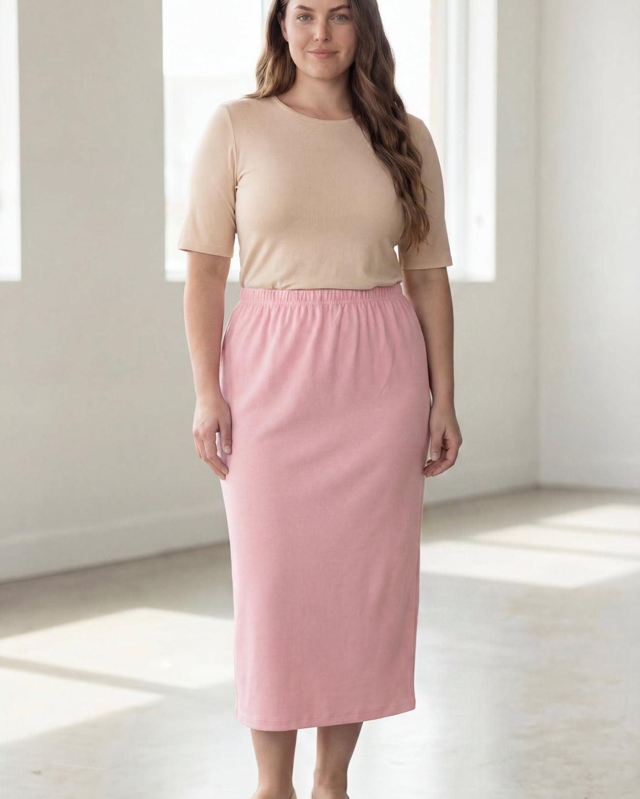 Woman wearing a beige shirt & pink maxi length pencil skirt standing in a bright room with large windows.