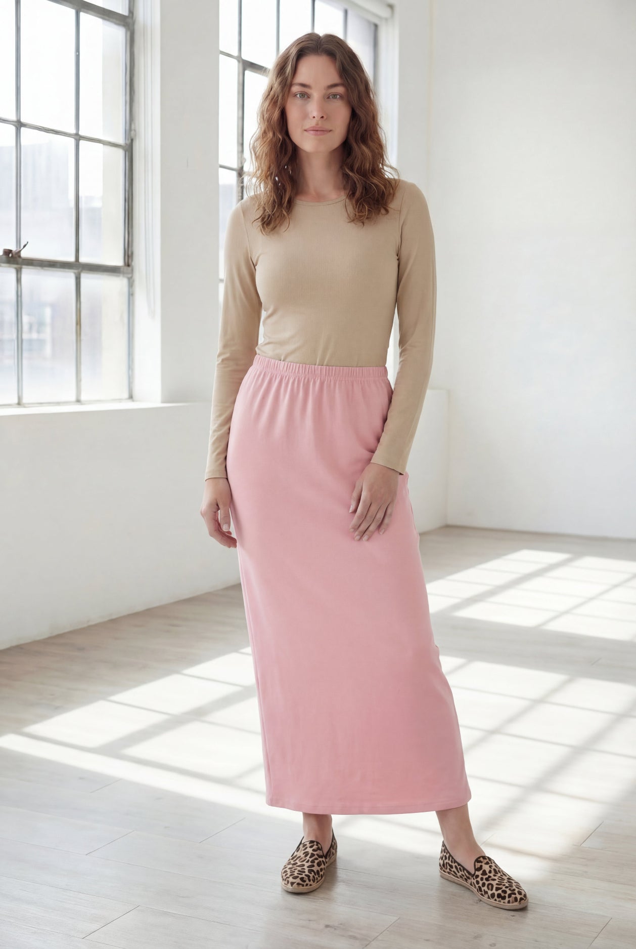 Woman wearing a beige shirt & pink maxi length pencil skirt standing in a bright room with large windows.