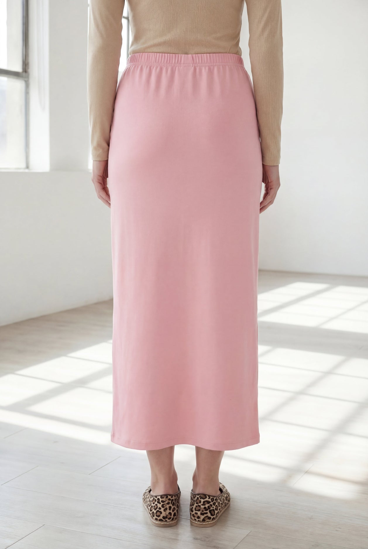 Woman wearing a beige shirt & pink maxi length pencil skirt standing in a bright room with large windows.