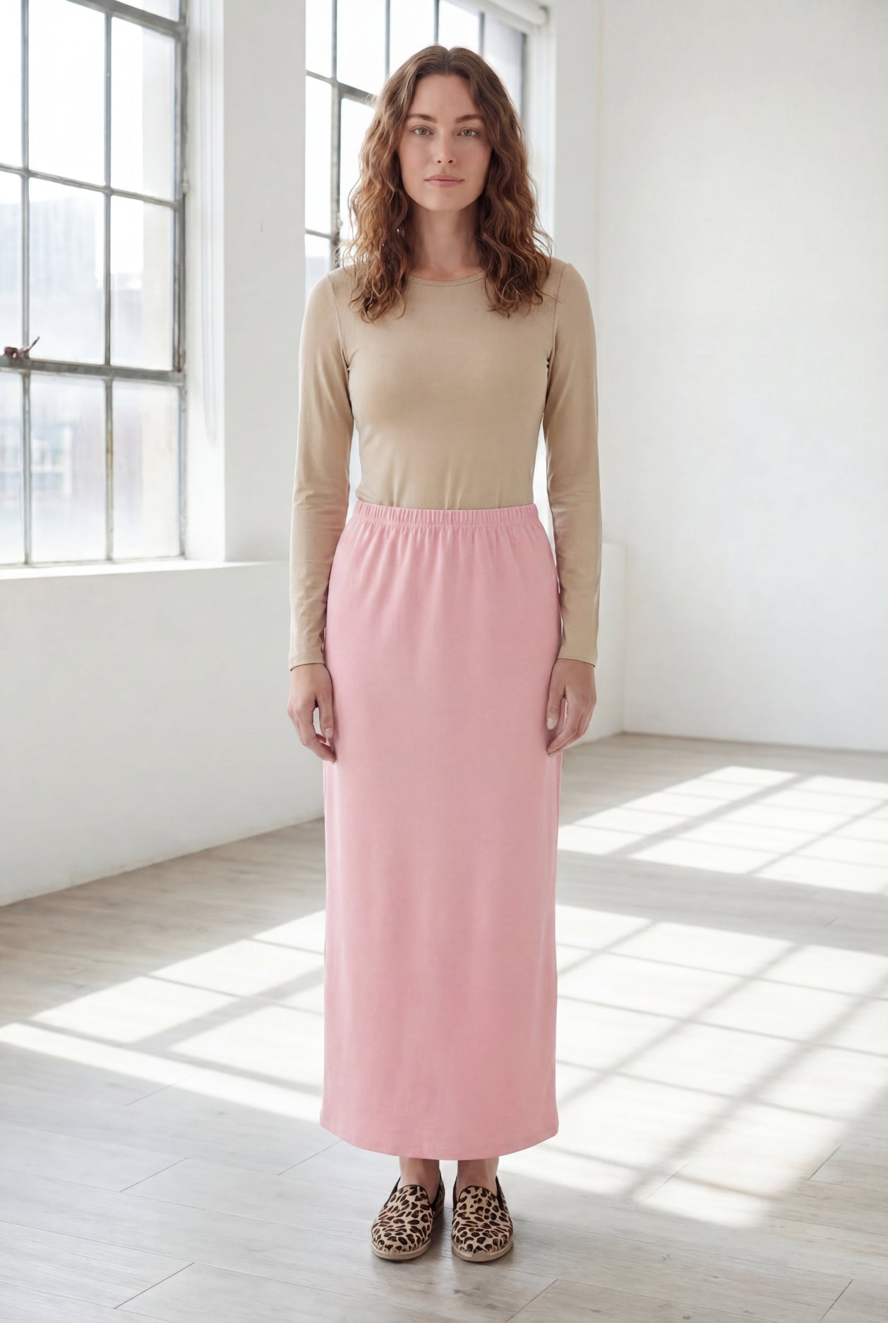 Woman wearing a beige shirt & pink maxi length pencil skirt standing in a bright room with large windows.