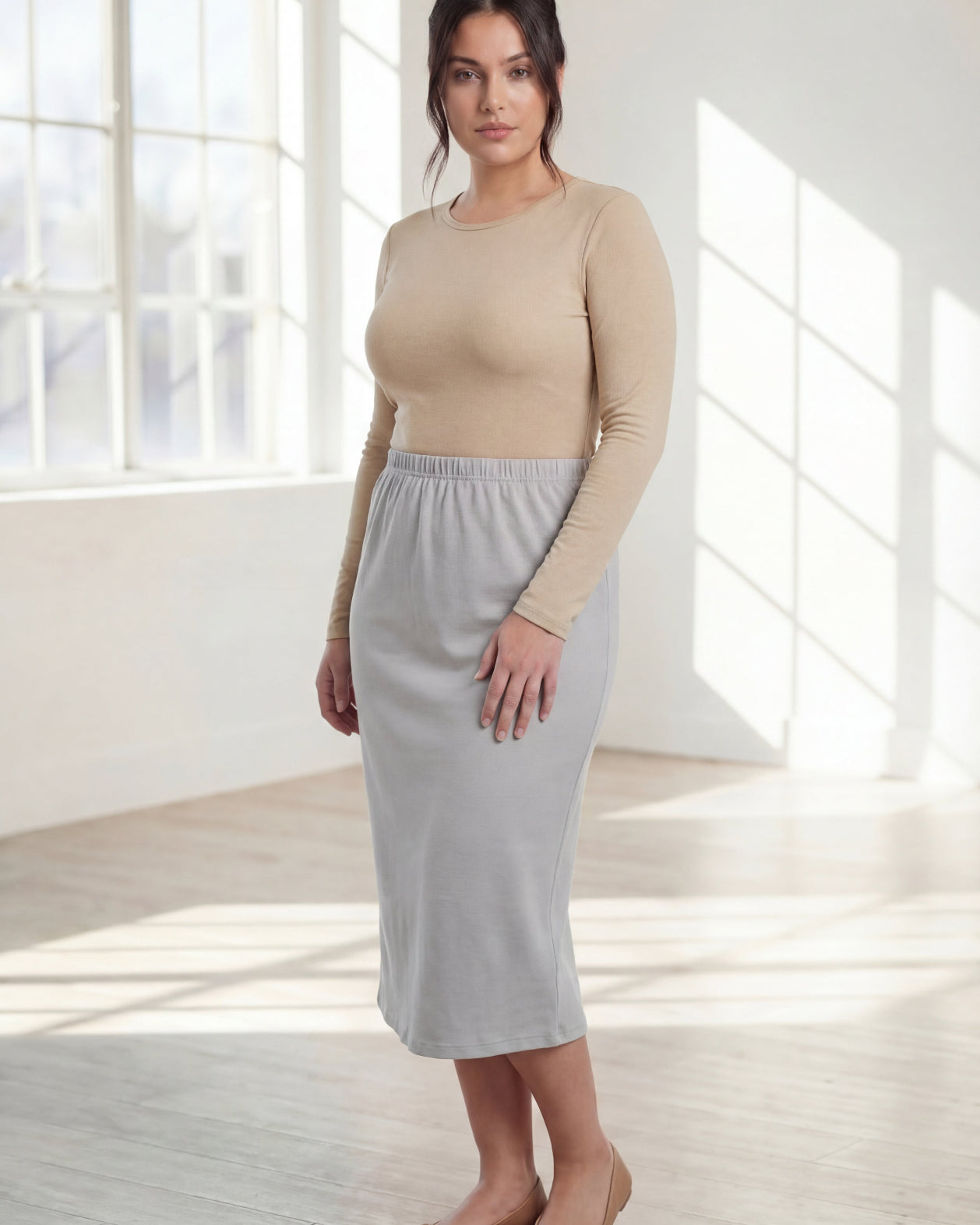 Woman wearing a beige top and silver skirt in a bright room with large windows.